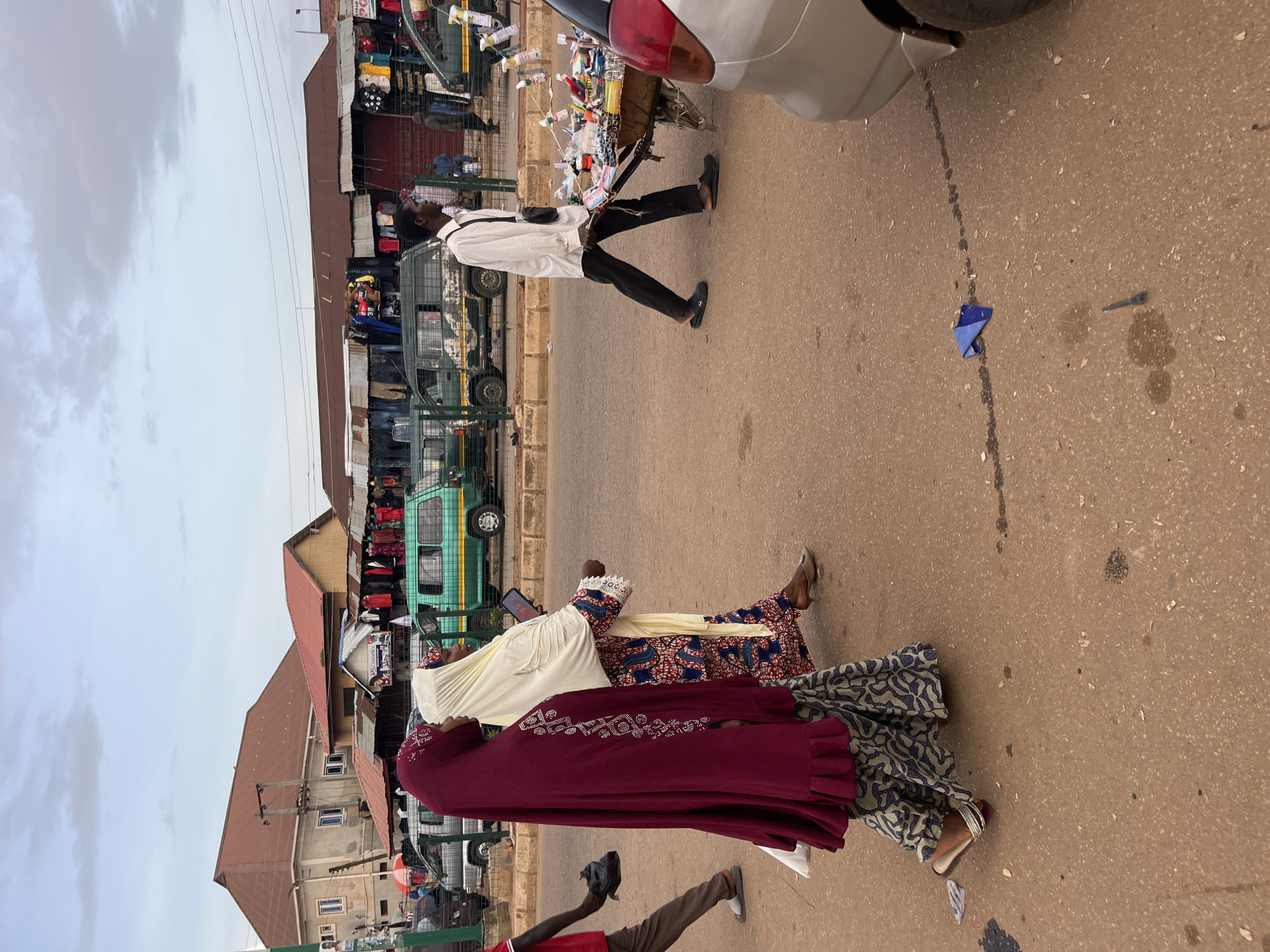 The image depicts a street scene in Bukuru, Nigeria, likely during daylight. The perspective is from a low angle, looking up at the subjects and the environment.
In the foreground, two women are walking away from the camera. The woman closer to the camera is wearing a dark red abaya adorned with intricate silver embellishments and a cream-colored headscarf. She is also wearing a patterned skirt with blue and white designs. The woman in front of her is wearing a similar patterned skirt and a white headscarf. Both women are wearing sandals.
Behind the women, a man in a white shirt and black trousers is walking towards the right side of the frame. He appears to be carrying a black bag slung across his chest. He is barefoot and wearing black slippers. He is walking past a cart filled with various items, possibly for sale. Further in the background, there are market stalls with clothing displayed, and several vehicles, including a green minibus. The buildings in the background suggest a bustling urban or semi-urban environment.
The ground is paved with asphalt. There are some scattered pieces of litter on the ground, including a crumpled blue paper and what appears to be a small plastic fork. A faint dark liquid stain is also visible on the asphalt. The sky above is overcast with clouds. The overall impression is a candid snapshot of daily life in a Nigerian market.