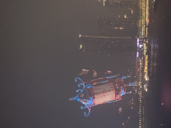 The image captures a nighttime urban scene in the 江北城街道 (Jiangbeicheng Subdistrict) of China. The primary subject is a large, elaborate drone light show formation depicting a traditional Chinese lantern, intricately outlined with red and blue lights, suspended against a dark sky. Several smaller, illuminated drone shapes, resembling butterflies or stylized birds, accompany the main lantern. Below the drone display, a body of water, likely a river, reflects ambient city lights. Along the far bank, a dense urban skyline is visible, characterized by numerous illuminated high-rise buildings and skyscrapers. A tall, slender structure with a vertical line of lights stands prominently on the right side of the cityscape. Several illuminated boats, likely passenger vessels, are present on the water near the illuminated riverbank. No individuals are discernible.