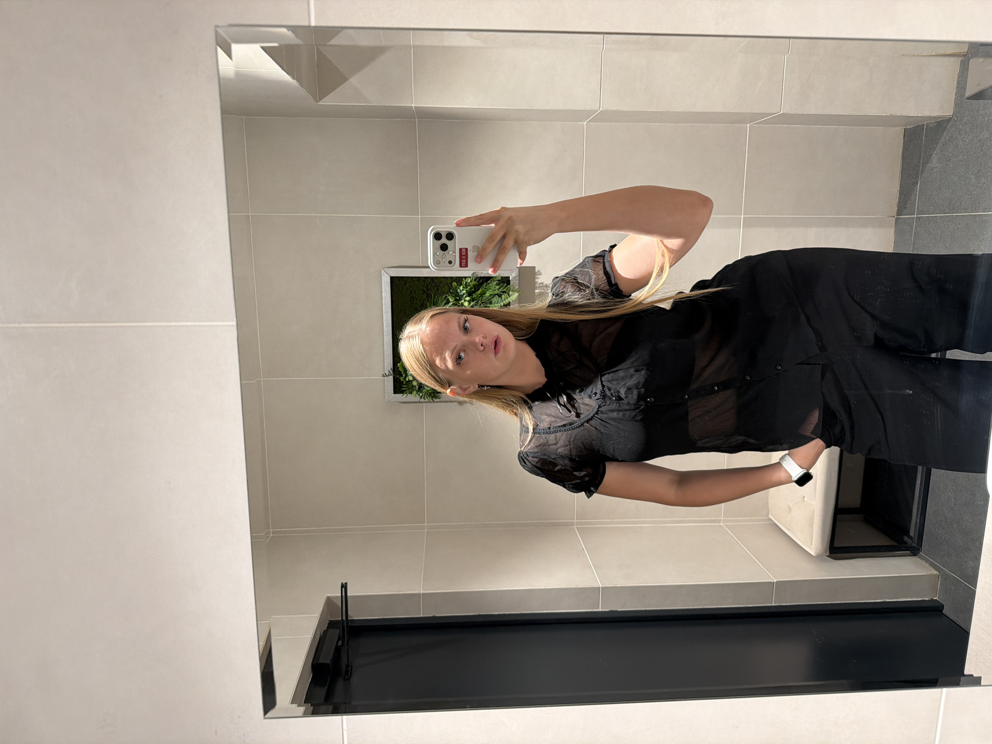 The media file shows a young woman taking a mirror selfie in what appears to be a modern bathroom. She has blonde hair and is wearing a black sheer blouse and black pants. She is holding a white smartphone in her right hand to take the photo. A watch with a white band is visible on her left wrist. The background consists of tiled walls in neutral tones and a black cabinet or fixture. A framed plant is visible in the reflection of the mirror behind her. The lighting suggests it is daytime, but no specific time of day can be determined. The overall mood is casual. There is some visible text on a sticker on the back of her phone case, which reads "KCK 9 GKH".