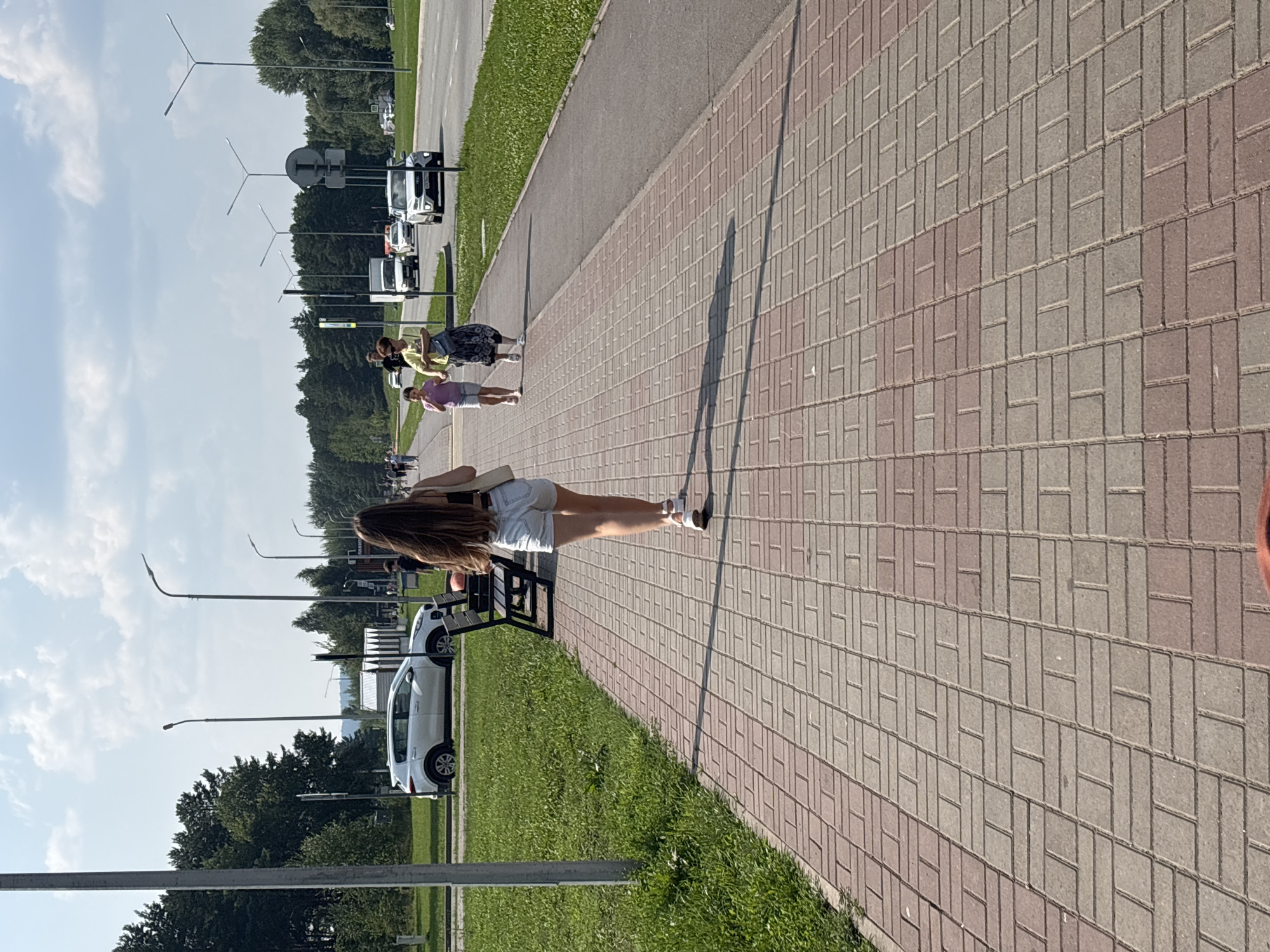 A full shot captures a woman from behind, walking away from the camera down a wide pedestrian walkway paved with red and grey bricks. She has long, dark hair and is wearing a black tank top and light denim shorts. Her right hand carries a light-colored tote bag. The walkway is bordered by a grassy area on the left and a road with moving vehicles on the right. Streetlights are visible along the walkway and the road. Further down the walkway, two other individuals, a woman and a young girl, are walking towards the camera. The woman is wearing a yellow shirt and a dark skirt, and the girl is wearing a pink top and white shorts. Trees line the background on the left, and a line of trees and buildings can be seen on the right side of the road. The sky is blue with scattered clouds. The scene appears to be a public space, likely in Naberezhnye Chelny, Russia, during daylight hours, possibly a park or a promenade adjacent to a street.