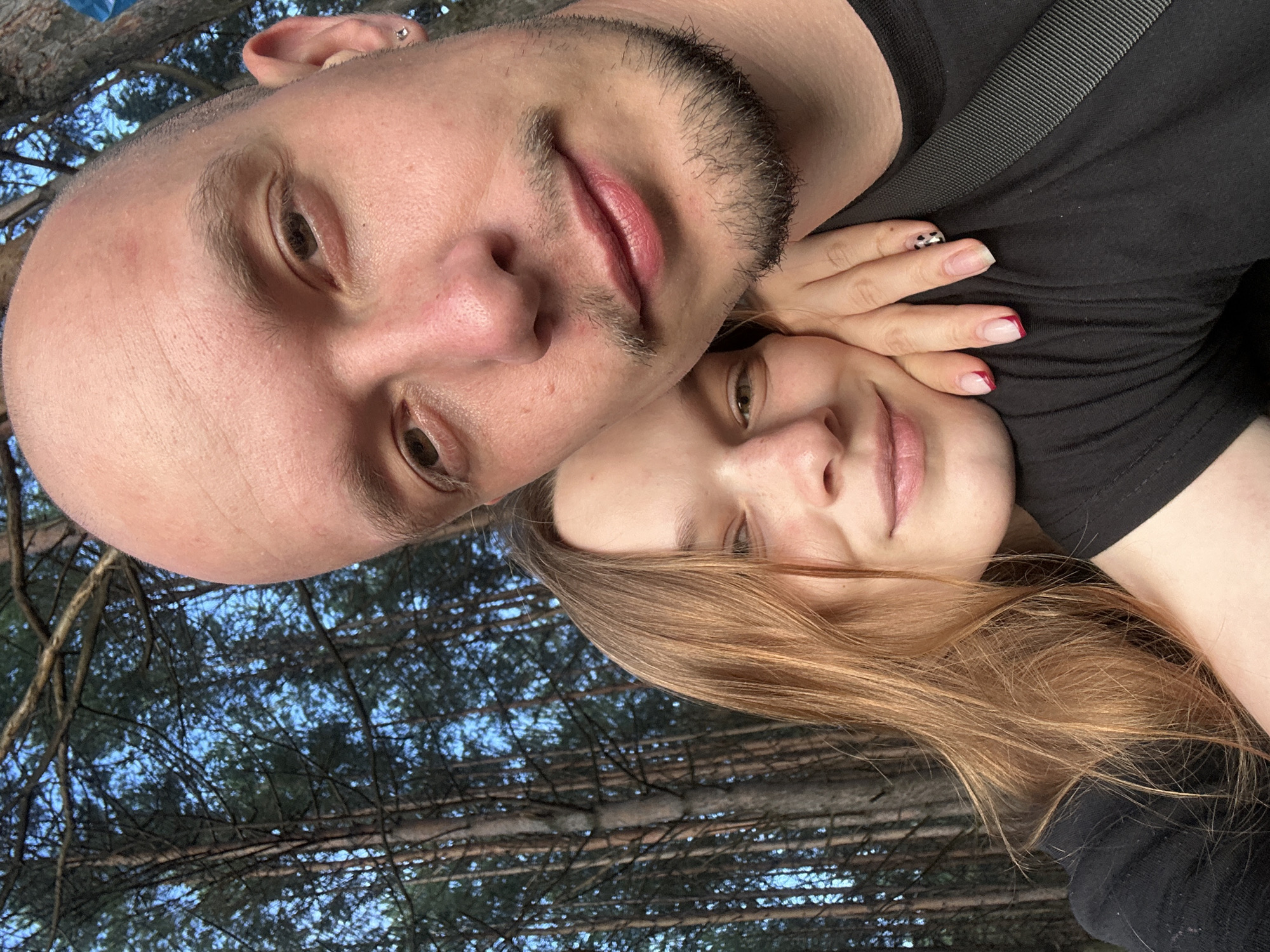 A close-up selfie shows a couple in a forest. The man, on the right, is bald with a short beard and mustache, and is wearing a black t-shirt. The woman, on the left, has long, reddish-brown hair and is leaning her head on his shoulder, her hand resting on his chest. Her fingernails have a French manicure with a red tip. The background consists of tall pine trees and a bright blue sky. The image appears to have been taken in or near Chaikovsky, Russia, given the presence of pine forests.