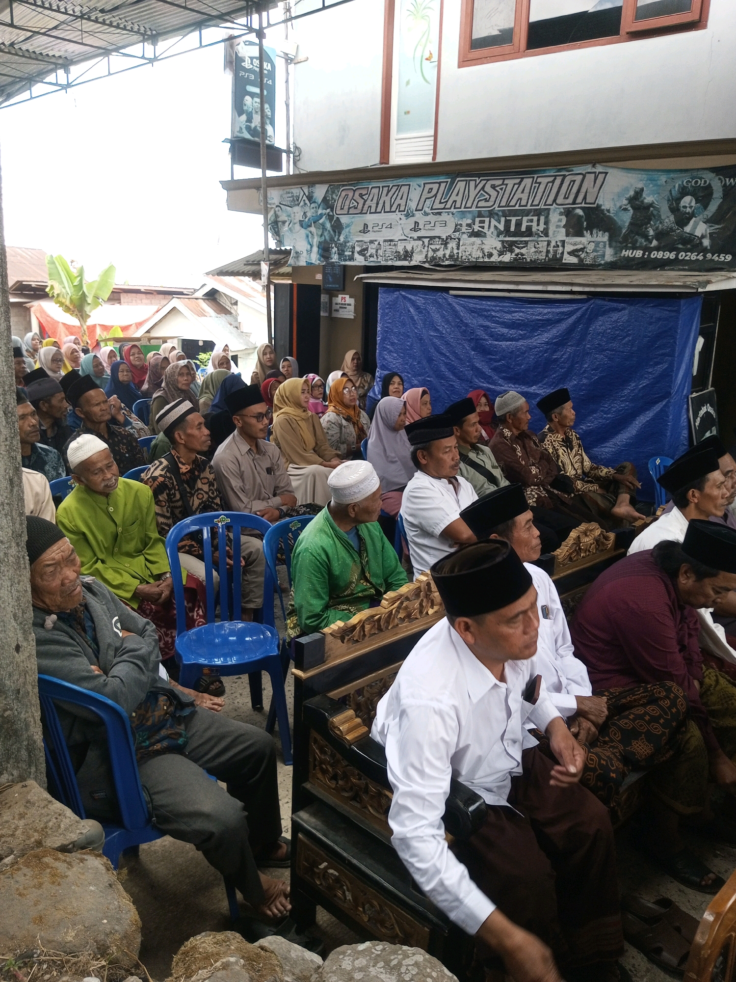 This image depicts an outdoor gathering of people, likely in the city of Andongsili, Indonesia, as indicated by the presence of a banner advertising "OSAKA PLAYSTATION" with Indonesian text and a phone number. The scene appears to be informal, with people seated on blue plastic chairs and ornate wooden benches. Many individuals are wearing traditional Islamic attire, including peci hats and hijabs, suggesting a religious or cultural event. The background features a building with a banner and a blue tarp, and some overhead structures that appear to be part of a temporary shelter. The lighting suggests it is daytime, with some overhead lighting visible. The overall impression is one of a community event or gathering.