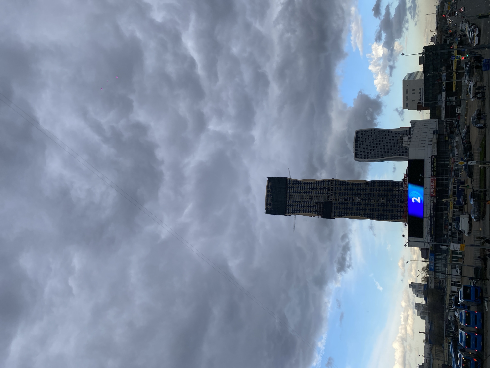 The image depicts a street scene in Moscow, Russia, under a sky dominated by storm clouds transitioning into blue sky. Two modern skyscrapers are visible, one under construction and displaying a large illuminated "2" on its facade. A second, white building stands nearby. Below the skyscrapers, there is a mix of commercial buildings, shops, and streetlights. A row of blue buses is parked along the curb. Cars are driving. The perspective is from an elevated viewpoint.