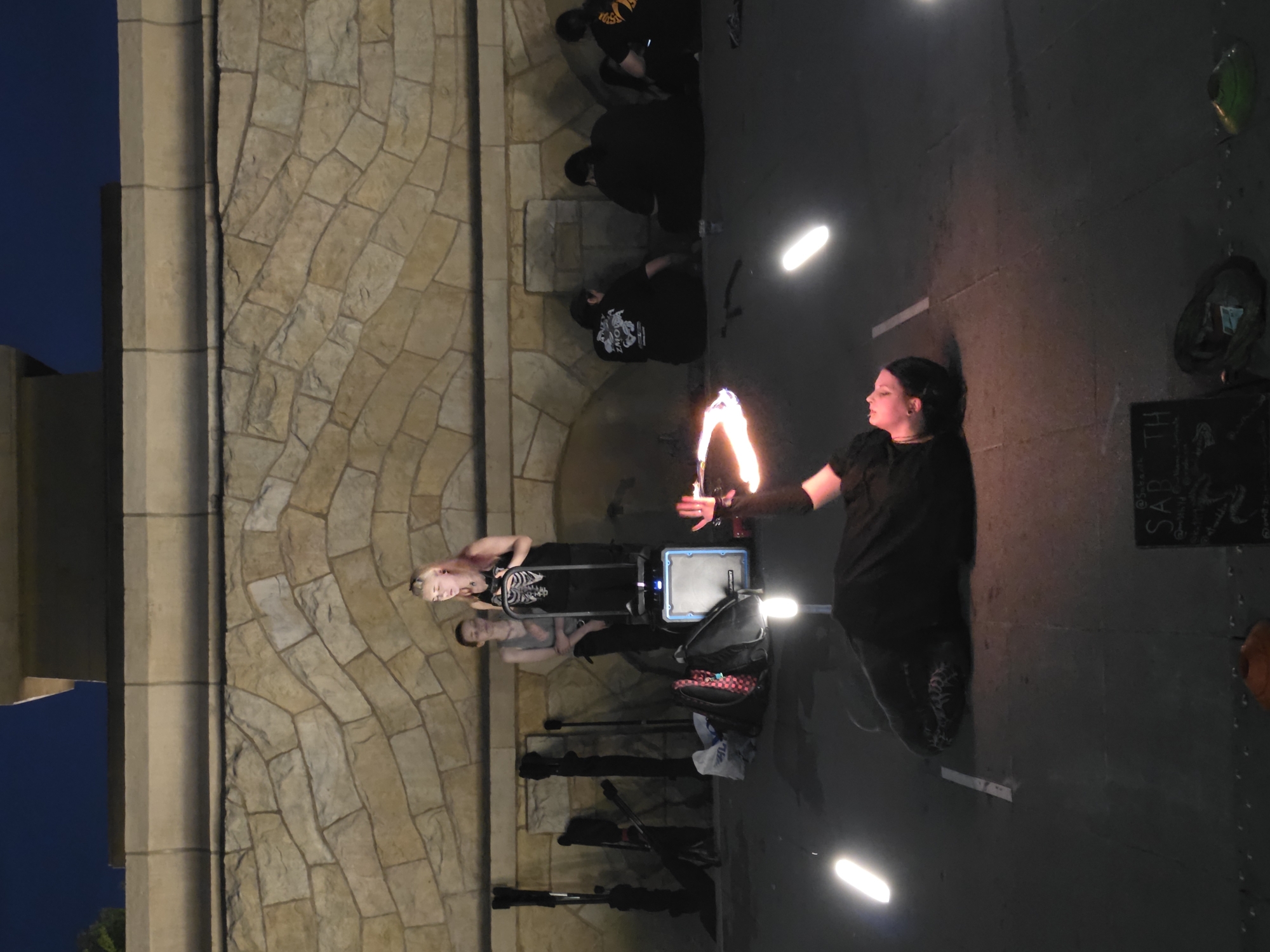 The image depicts an outdoor performance in Warsaw, Poland, at night. In the foreground, a person lies on their back on a paved surface, performing with fire. Their right hand is raised, and a ring of fire is visible around it. Several bright, elongated lights are positioned around the performer, casting a glow on the ground. In the mid-ground, two individuals stand behind a portable speaker. One is wearing a black top with a skeleton design, and the other wears a grey tank top. Various props, including bags and poles, are scattered on the ground behind them. The background features a textured stone wall and the dark night sky. Further back, several other individuals are seated on the ground, some wearing dark t-shirts. A small chalkboard with the word "SARTH" is visible in the lower right corner of the frame. The overall scene suggests a street performance or outdoor event.