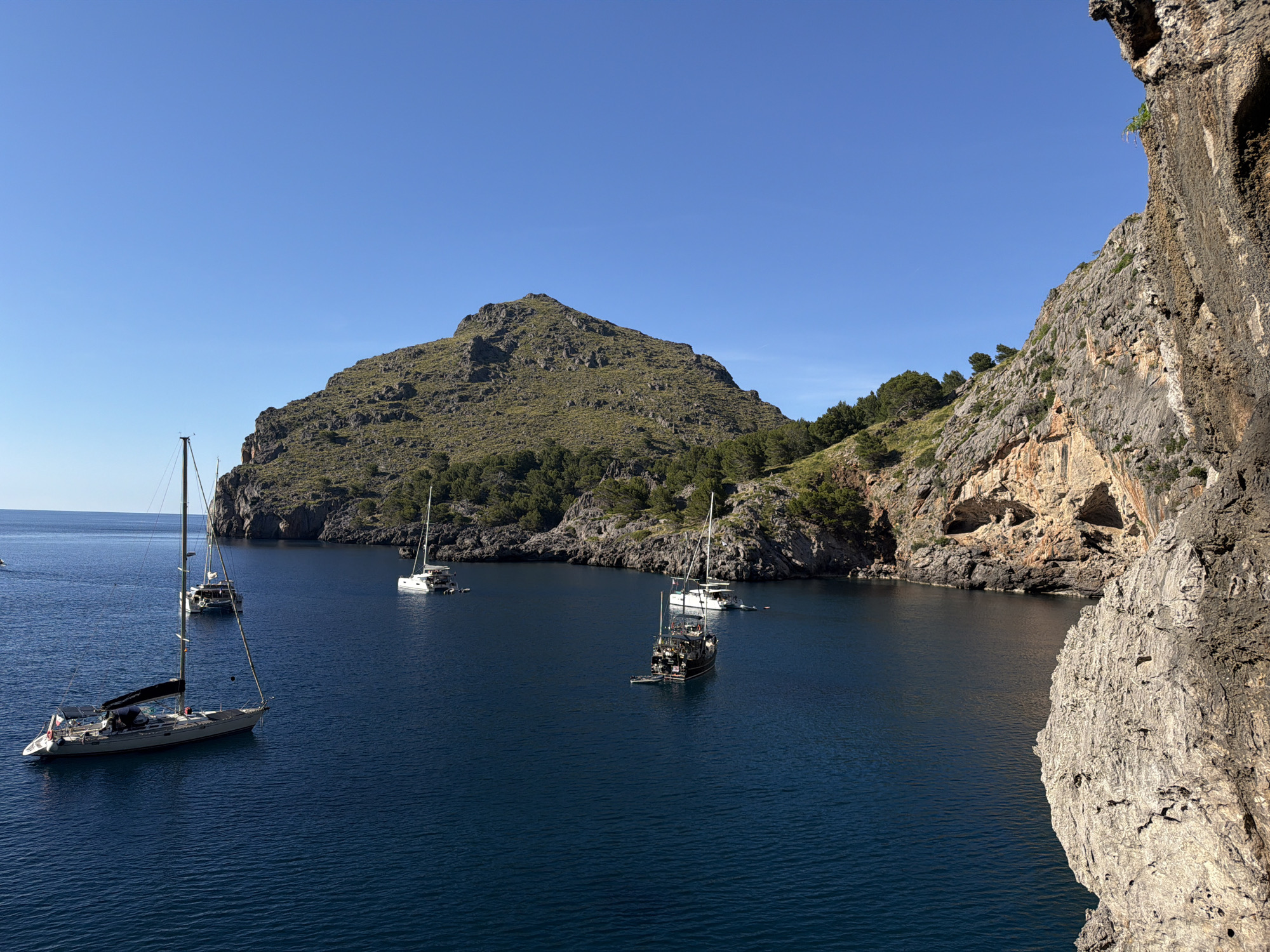 The image depicts a coastal inlet near Escorca, Spain, filled with deep blue water. Several sailboats and other watercraft are anchored in the bay. A large, mountainous landmass, covered with vegetation, forms a prominent feature in the background. To the right, a tall, rocky cliff dominates the foreground, showing various geological textures and sparse vegetation. The sky is clear and blue.