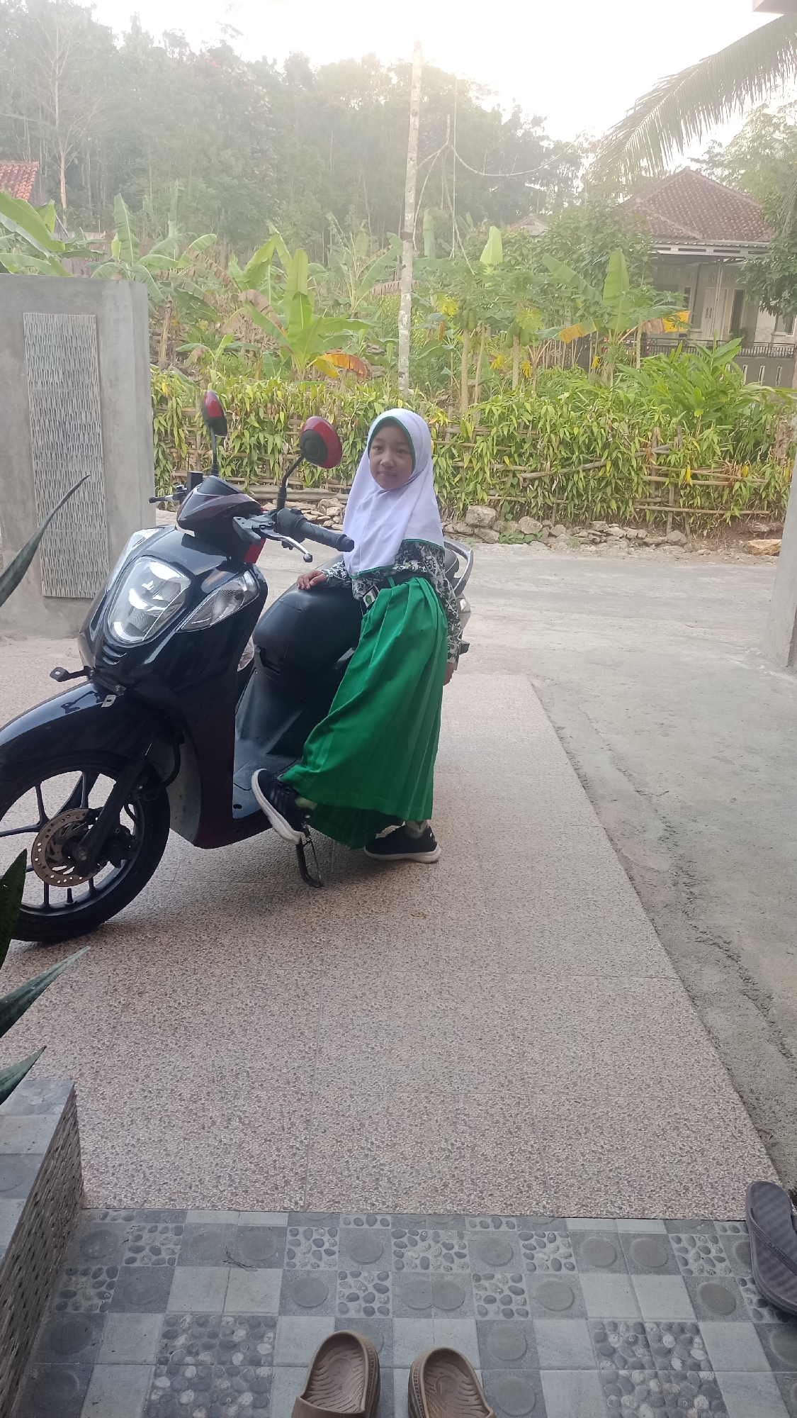A young girl wearing a white hijab and a green skirt sits on a black and maroon scooter. She is on a paved walkway in front of a building. In the background, there are trees and a house with a red roof. The scene appears to be in a rural or suburban area, likely Pakemitan, Indonesia, given the vegetation and style of housing.
