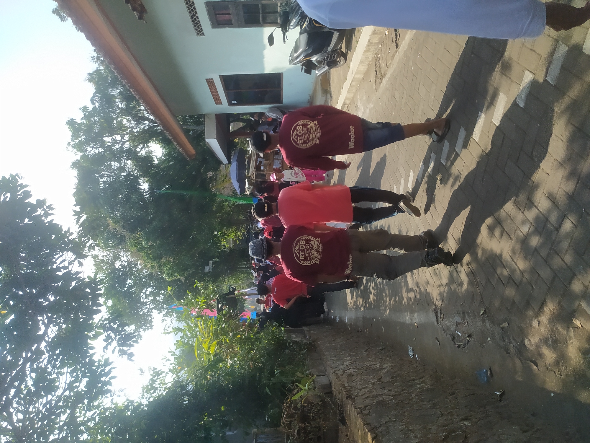 A group of people walk along a paved path next to a low stone wall. The path is lined with trees and a light-colored building with a tiled roof. The building has several windows and a parked scooter in front of it. The people are dressed in casual clothing, with some wearing red shirts with logos. The sun is shining, casting shadows on the ground. Some of the people appear to be walking in a procession or event. The scene is located in the city of Lopait, Indonesia.