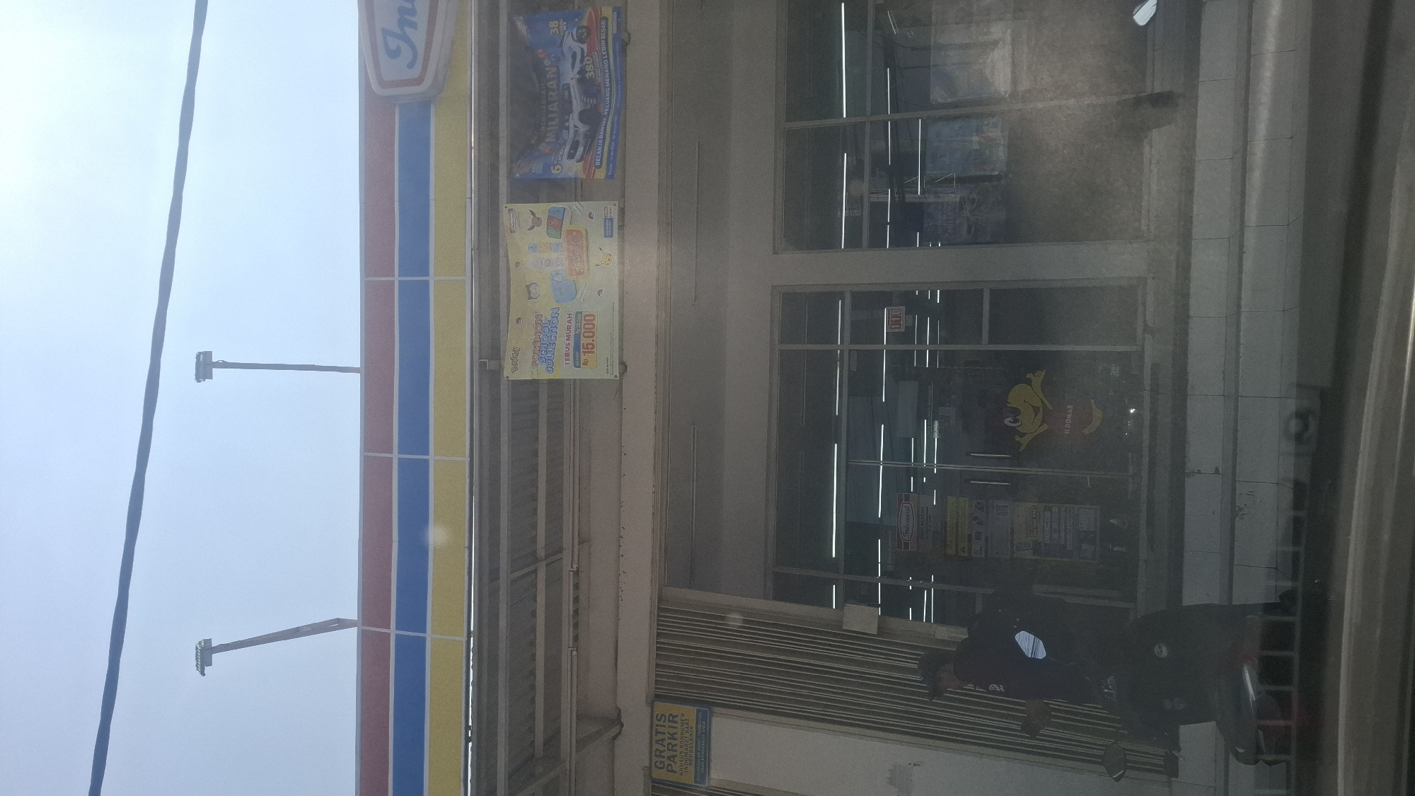 The image captures the exterior of an Indomaret convenience store in Rancagoong, Indonesia, under a bright, overcast sky. The store's distinctive red, blue, and yellow striped awning is visible across the top of the building. A promotional banner featuring "Pokemon School Collection" with a price of Rp 15.000 is displayed on the facade. Another banner, predominantly blue, shows a car and text mentioning "MILIARAN," suggesting a lottery or prize promotion.
In front of the store, a metal gate is partially open, revealing a glimpse of the interior. A person wearing a dark uniform, possibly a store employee, is standing near the entrance, holding a mobile phone. A motorcycle is parked in front of the gate. Inside the store, through the glass doors, rows of fluorescent lights illuminate the shelves. A cartoonish yellow character with red accents, possibly a mascot, is visible on the glass door. A sign that reads "Indomaret" is also present on the store's entrance. A blue sign on the left side of the building states "GRATIS PARKIR KHUSUS KONSUMEN INDOMARET" indicating free parking for Indomaret customers. The overall environment suggests a typical street view of a commercial area.
