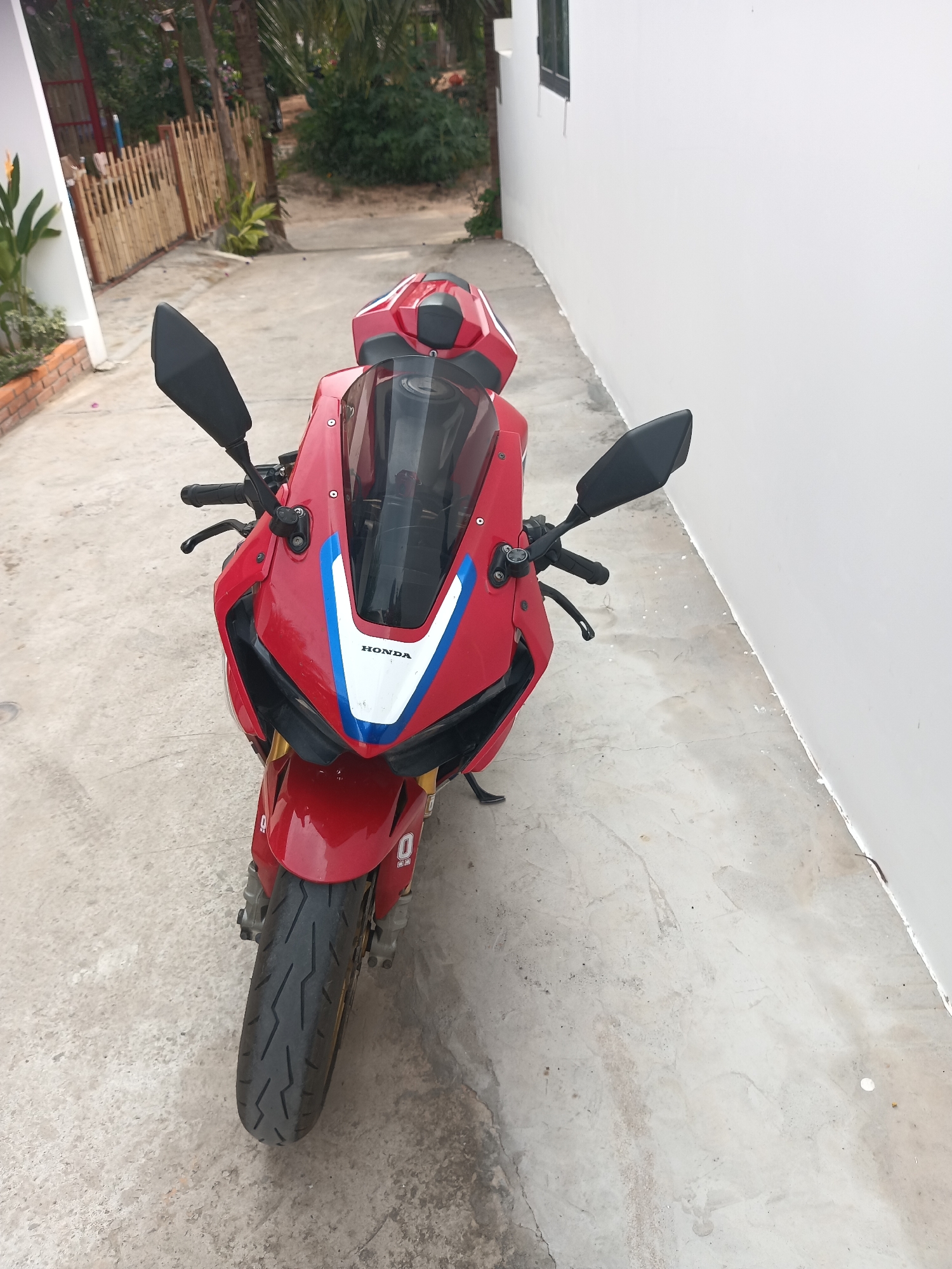 A vibrant red sport motorcycle, identified as a Honda by the "HONDA" branding on its front fairing, is parked centrally in this outdoor scene. The motorcycle features white and blue graphic accents on its fairing, a dark windscreen, black mirrors, and gold-colored front fork components. Small '0' logos are visible on the front fender and lower fairing.
The motorcycle is resting on a light grey concrete or paved surface, which shows signs of wear, minor cracks, and scattered dirt. To the left of the motorcycle, a low brick planter contains green foliage, beyond which a rustic bamboo fence runs alongside a path. Tropical greenery, including palm trees and various bushes, fills the area behind the fence, suggesting a lush, warm climate. To the right of the motorcycle, a tall, unadorned white wall of a building stretches into the background, with a dark-framed window positioned higher up the wall.
The lighting is bright and diffused, indicating it is daytime, possibly under an overcast sky, as there are no harsh shadows. No activity is taking place; the motorcycle is stationary. The setting, with its tropical plants and building style, is consistent with a location such as Mũi Né, Vietnam.