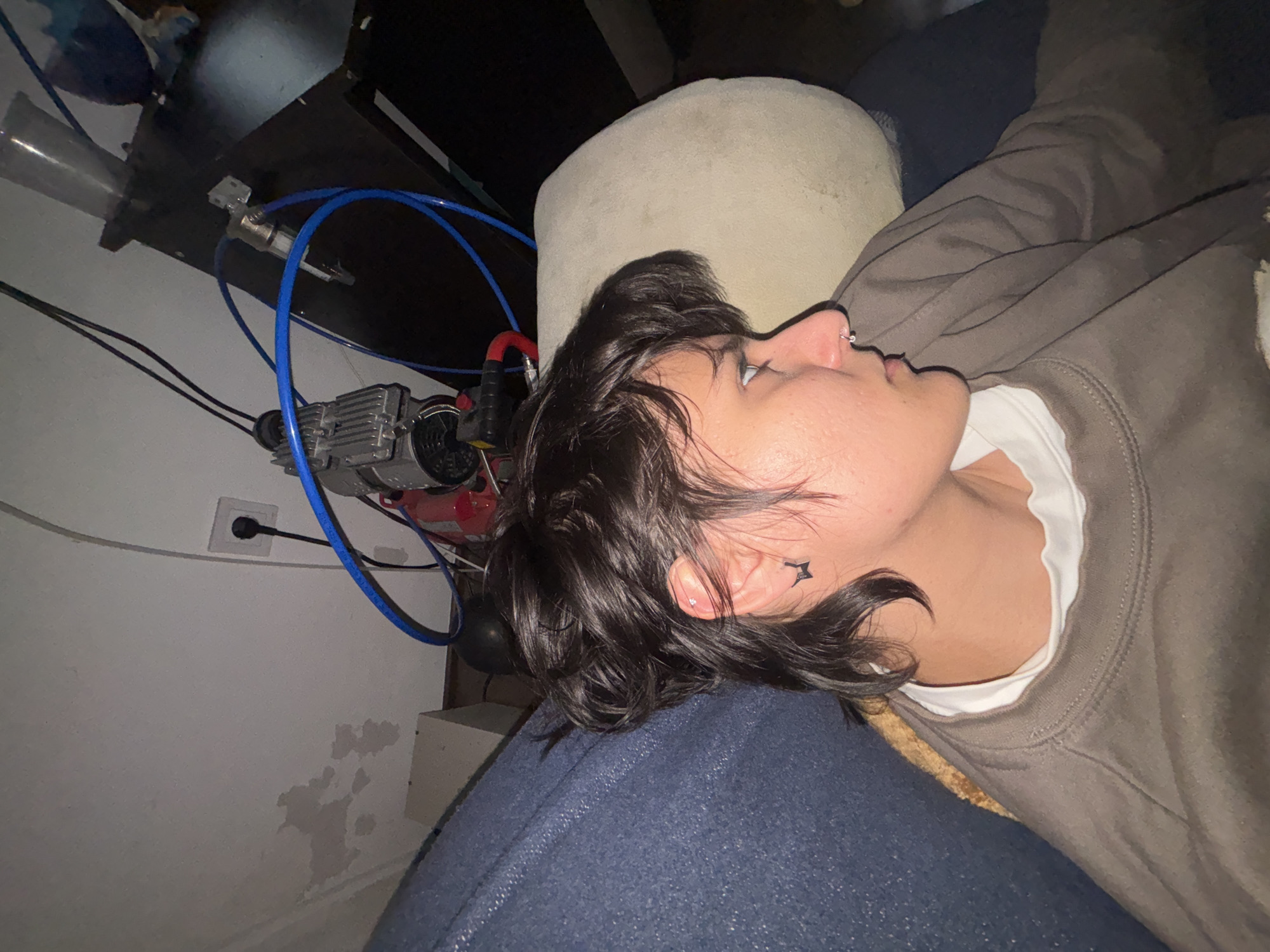 The image depicts a person lying on a blue couch, resting their head on the couch's arm. They are wearing a brown sweatshirt over a white undershirt. The person has dark hair and a star tattoo near their left ear, which is also pierced. Visible behind the person are a light-colored cushion, a wall with an electrical outlet, and a setup of blue tubing connected to a pump or similar device. There is also a small black table next to the couch. There is no indication the image was taken in Sofia, Bulgaria.