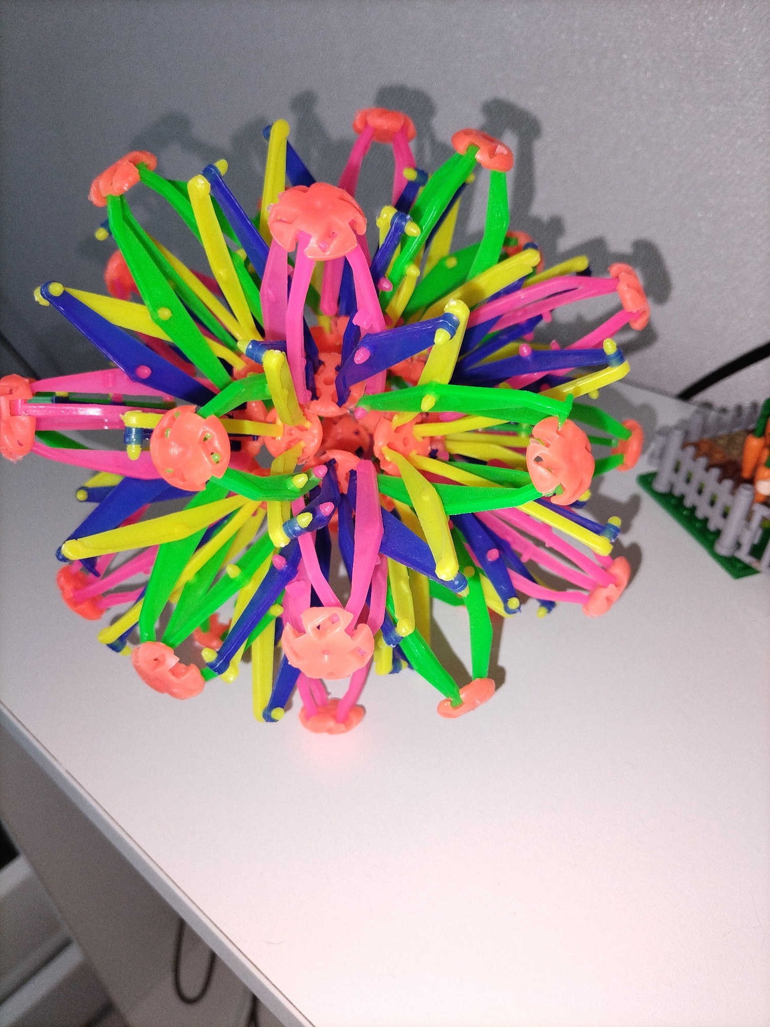 This media file contains a close-up, top-down view of a brightly colored, collapsible ball toy. The toy is composed of numerous interconnected plastic rods in vibrant colors such as pink, yellow, green, and blue. At the ends of many of these rods are small, orange, flower-shaped connectors that allow the structure to expand and contract. The ball is positioned on a plain white surface, likely a table or shelf, with a neutral gray background behind it. In the upper right corner of the image, a small portion of another toy is visible, featuring a white picket fence and what appear to be miniature carrots, suggesting a collection of toys. The lighting in the image casts shadows from the toy onto the background, indicating an indoor setting, possibly during daytime or with artificial lighting. There is no text visible in the image. The focus is entirely on the intricate and colorful construction of the expandable ball.