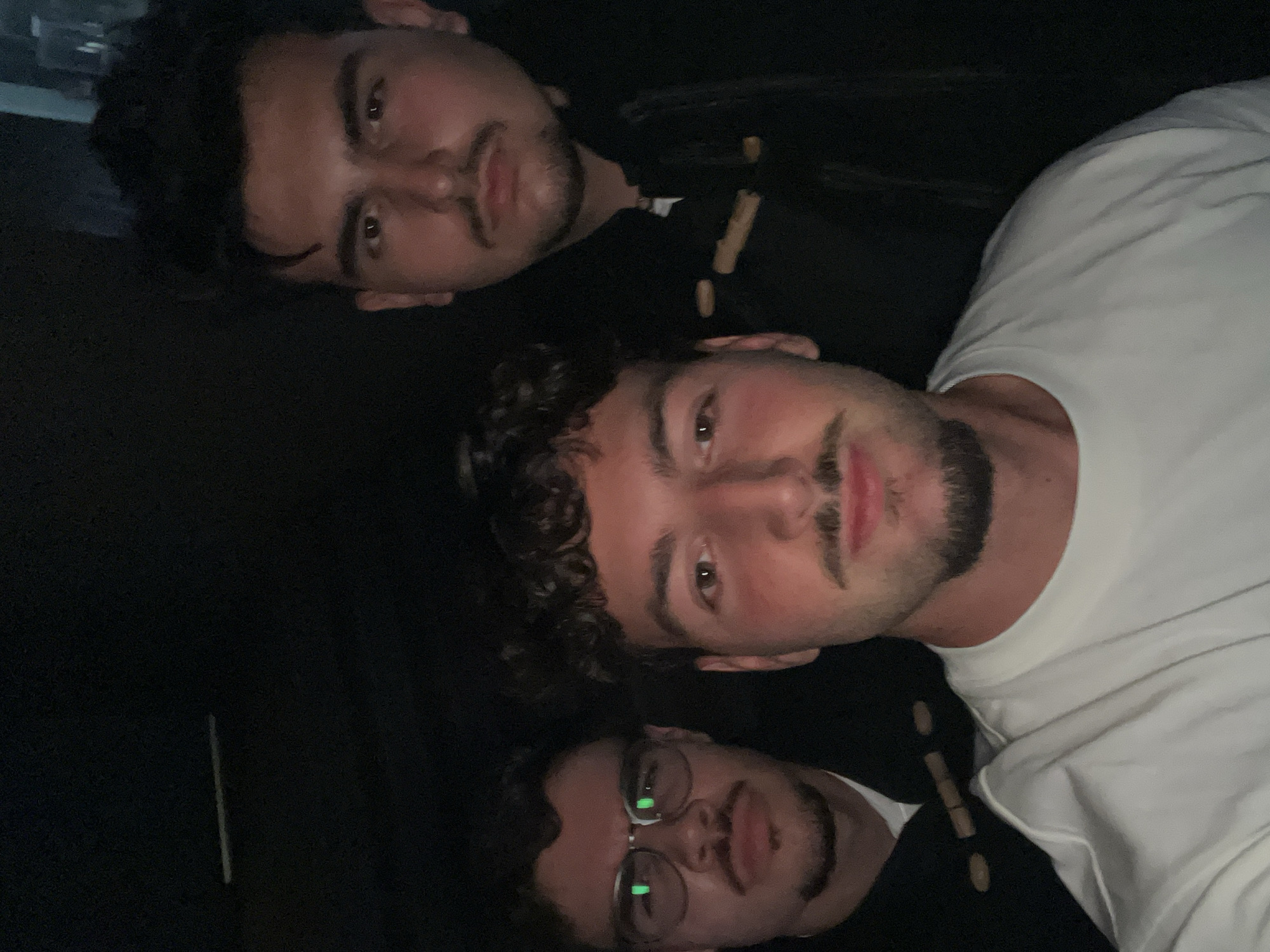 The image captures a casual, close-up group photo of three young adult males, presented in a portrait orientation (rotated 90 degrees counter-clockwise from a standard upright view). The scene is dimly lit, suggesting an indoor setting or nighttime.
From top to bottom in the image's current orientation:
The topmost individual has dark, curly hair and a mustache with a short beard. He is wearing a white t-shirt. His gaze is directed towards the camera with a neutral expression.
Below him is another male, also with dark, curly hair and a short beard, wearing a dark top. He is looking at the camera with a serious or neutral expression.
At the bottom, partially visible, is a third male with dark hair, wearing glasses, and also sporting a mustache and short beard. He is wearing a dark top, and reflections are visible on his glasses.
All three subjects have light to medium skin tones. Their faces fill a significant portion of the frame, indicating a close-up shot, possibly a selfie. The background is uniformly dark, offering no specific details about the environment beyond the low light. Small, elongated brown objects, possibly beads or decorative elements, are scattered on the dark fabric visible between the subjects.
The activity is a simple posed group photo. No specific event is discernible, but the direct gazes imply an awareness of being photographed. There is no visible text. The location context provided is Vila Real, Portugal, though no visual cues within the image confirm this. The overall mood is casual and relaxed.
