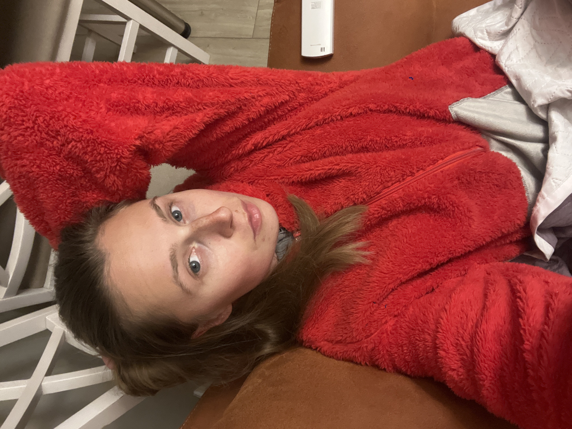 A young woman with fair skin and blue eyes is lying on what appears to be a brown couch or cushioned surface. She is wearing a bright red, furry zip-up jacket. Part of a white crib or bed frame is visible to her left. There is also a small white electronic device in the background. The location is not discernible from the image alone.