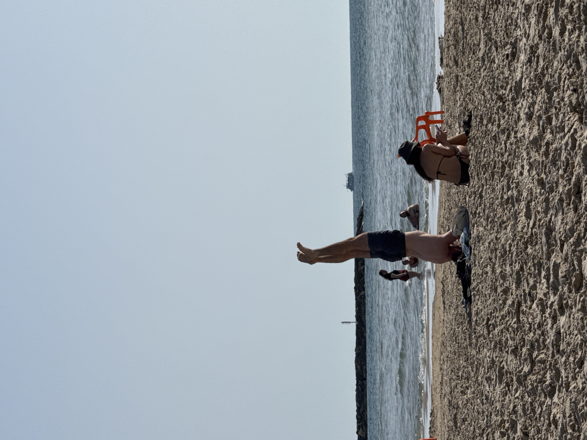 The image shows a beach scene in Tel-Aviv, Israel, oriented with the sky on the left and the sandy beach on the right. The sea runs in a vertical strip between the sky and the sand.
Several people are present on the beach. A woman in a black bikini and hat sits on the sand near an orange plastic chair. Nearby, a man is lying on the beach. Another person is standing near the water's edge. A boat is visible in the sea.