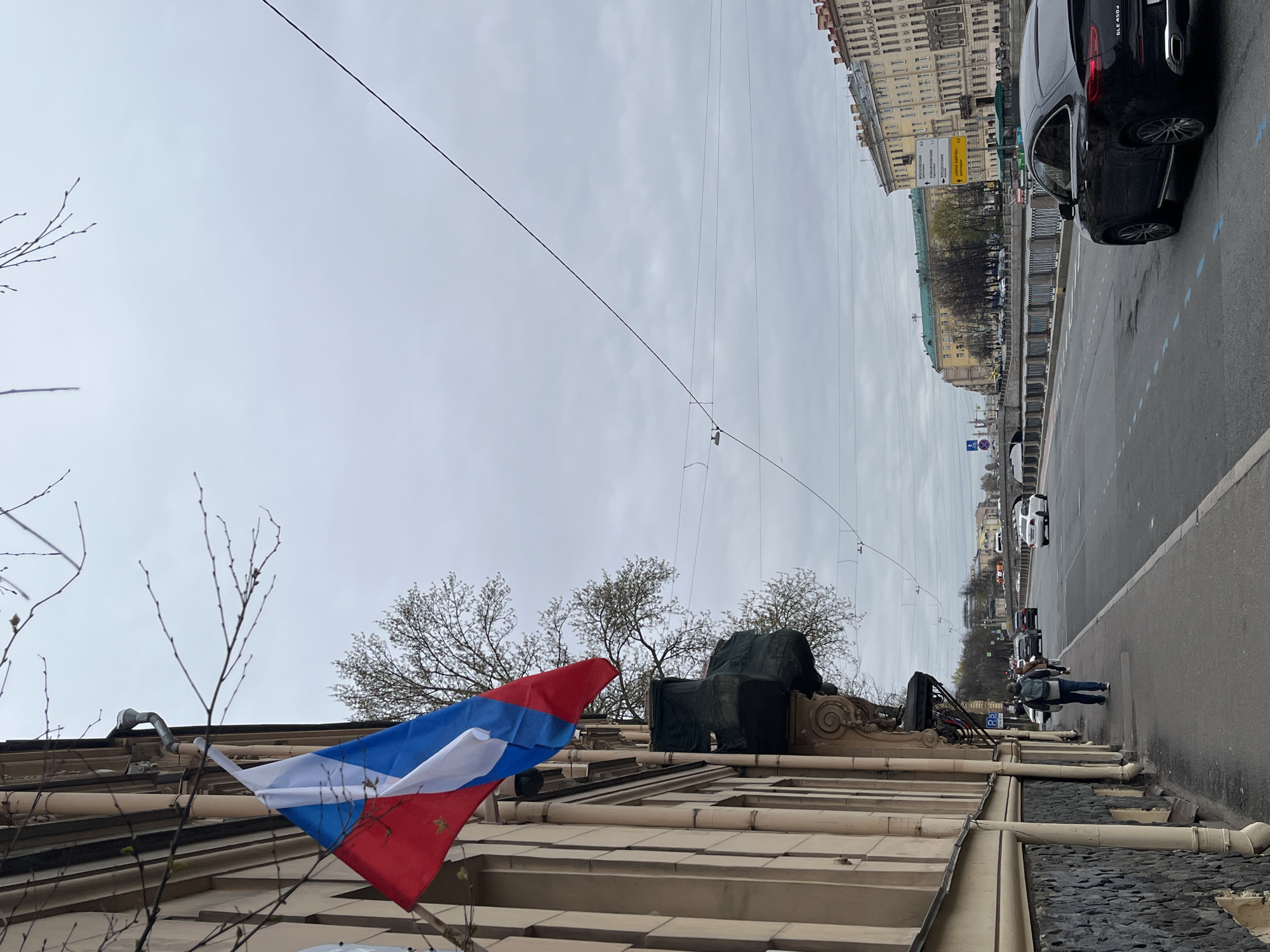 The image, viewed horizontally, depicts an urban street scene on an overcast day. In the foreground on the left, the facade of a light-colored building with ornate architectural details is visible. A Russian flag (white, blue, red horizontal stripes) hangs from this building. On the building's roof, a large, dark, draped object, possibly a statue, stands next to budding tree branches.
The midground features a street or embankment with a sidewalk. Several pedestrians walk along the sidewalk, and cars are parked along the curb, including a prominent black SUV (Mercedes-Benz GLE 350d) with its rear visible. White dashed lines mark the road.
In the background, across the street, are multi-story buildings with classical architecture, some featuring green roofs. Overhead, numerous power lines crisscross the grey, cloudy sky. A yellow street sign with Cyrillic text is clearly visible on the far side of the street, indicating directions to "МОСКОВСКИЙ ПРОСПЕКТ" (Moskovsky Prospekt), "ИЗМАЙЛОВСКИЙ ПРОСПЕКТ" (Izmaylovsky Prospekt), "КРЮКОВ КАНАЛ" (Kryukov Canal), and "МОСТ КРАСНОАРМЕЙСКИЙ" (Krasnoarmeysky Bridge), strongly suggesting the location is Saint Petersburg, Russia. A blue "P" parking sign is also present. The scene suggests a busy city environment during daytime, possibly in early spring given the budding trees.