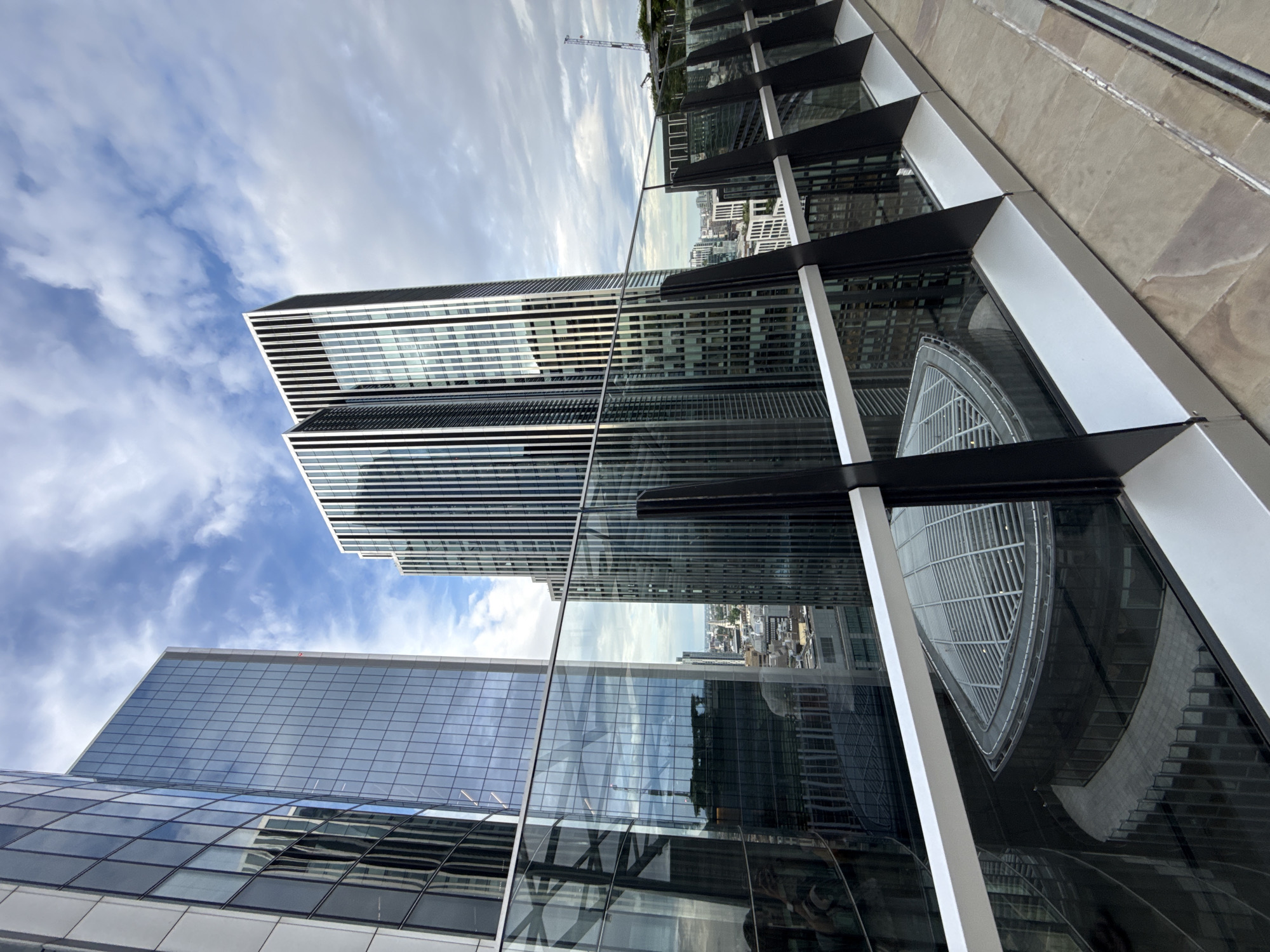 This image captures a view of modern architecture, likely in London, United Kingdom. The primary subjects are several contemporary skyscrapers with glass facades. One prominent building features horizontal white stripes against a dark background, creating a distinct visual pattern. Another building displays a large, curved glass structure, reflecting the sky and other buildings.
The scene is set on a clear day with a partly cloudy sky, suggested by the white clouds against the blue. The reflections in the glass surfaces indicate a daytime setting, and the sharp shadows cast by the buildings further support this. The perspective is from a high vantage point, possibly another building or a rooftop, looking down and across at the surrounding urban landscape. The angle of the shot is tilted, giving a dynamic and slightly abstract feel to the composition.
Notable details include the intricate patterns of the building exteriors and the complex reflections. The presence of a construction crane in the background hints at ongoing development in the city. The overall atmosphere is one of urban dynamism and architectural innovation. No visible text is present in the image.