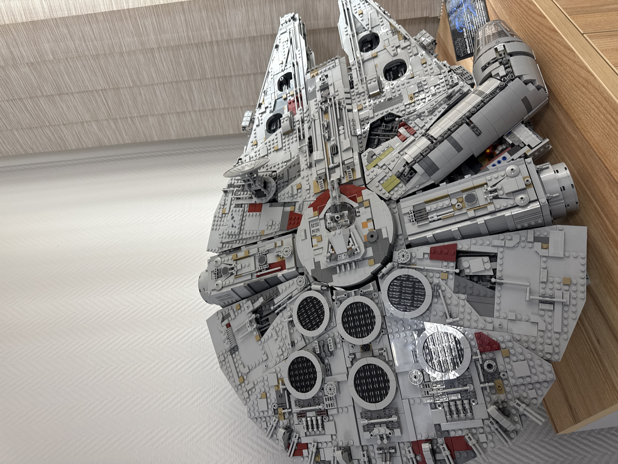 A high-angle, close-up shot depicts a large, assembled Lego Millennium Falcon model. The model is predominantly grey and white, with red accents. It is positioned against a textured white wall on the left and a wooden surface on the right. A Lego Star Wars box is visible in the background. The model is detailed and appears to be resting on a surface.