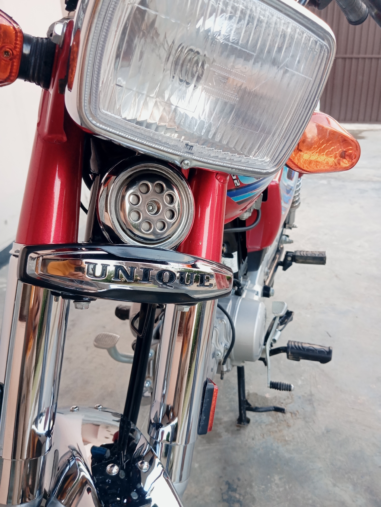 A close-up, eye-level, slightly angled shot captures the front of a red and chrome motorcycle. The focus is on the front fork, headlight, and a prominent chrome emblem that reads "UNIQUE." The motorcycle has a classic design, with a large, clear headlight and amber turn signals. The chrome elements are highly reflective, mirroring the surroundings. In the background, a light-colored wall and a brown fence are visible, suggesting an outdoor setting, possibly a driveway or parking area. The ground appears to be concrete. There are no people or animals present.