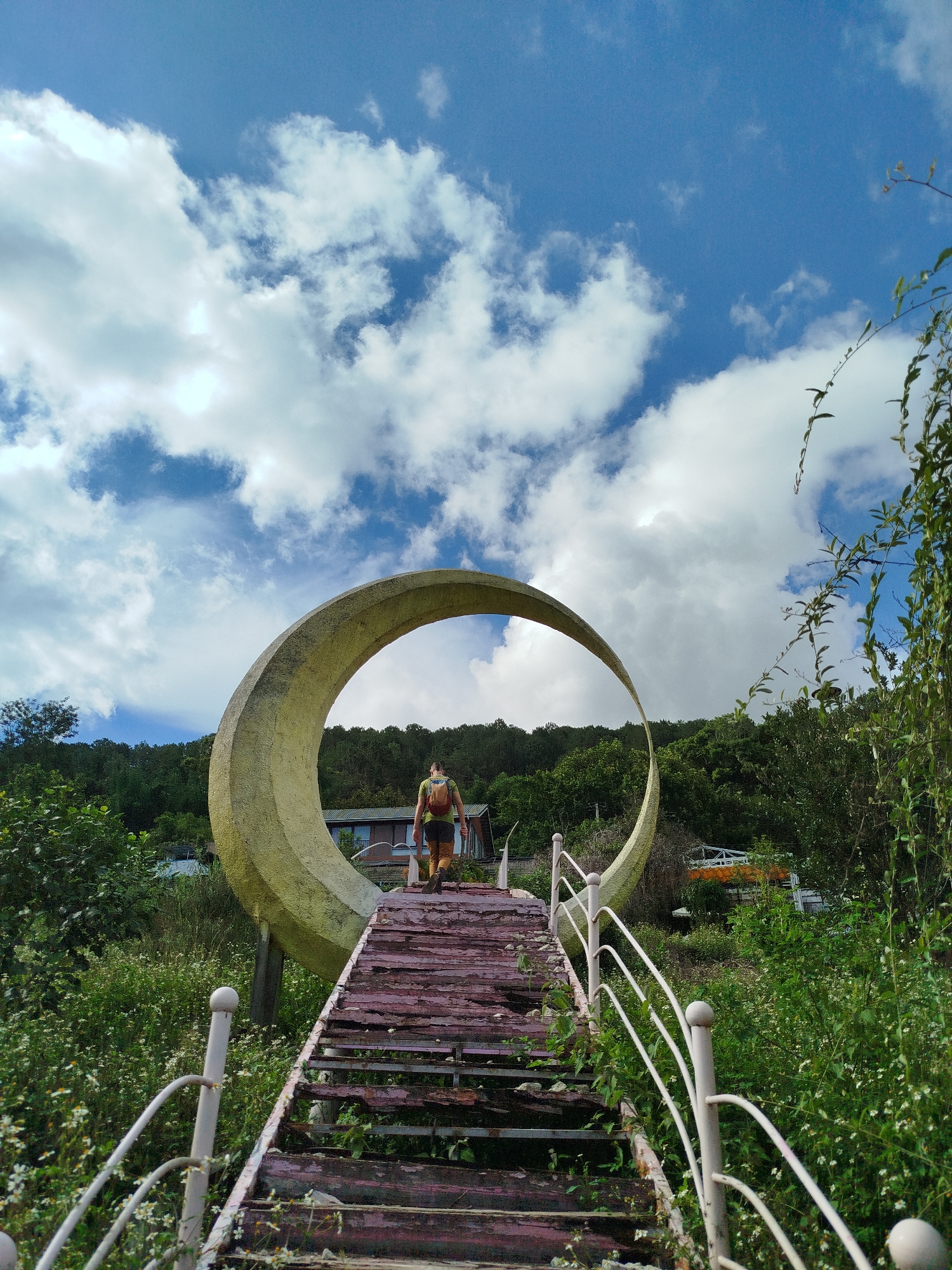 A person walks up a set of deteriorating wooden stairs with a white metal railing towards a large, crescent-shaped sculpture under a blue sky with scattered white clouds. The location is likely Da Lat, Vietnam. The person is wearing a backpack and is positioned near the top of the stairs, passing through the crescent. Behind the crescent, green trees and a building are visible. The stairs and surrounding area are overgrown with greenery and small white flowers.