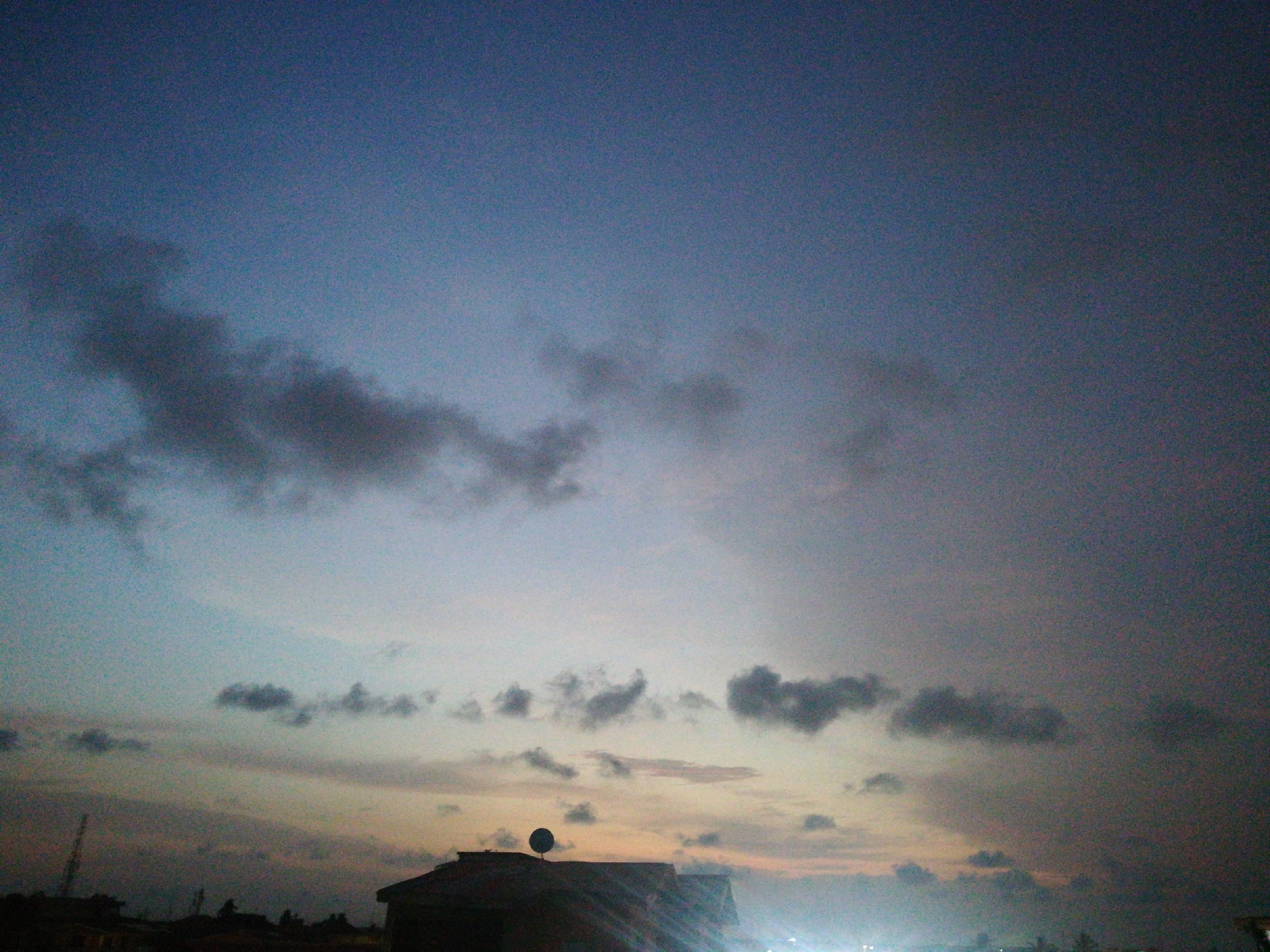 The image depicts a cityscape at dusk or dawn, likely in Lagos, Nigeria, based on the presence of a communication tower and architectural styles. The sky is a gradient of blue, gray, and orange hues, with scattered dark clouds. In the foreground, there are silhouettes of buildings, one with a satellite dish, and other low-lying structures. There is also light, possibly from streetlights or other buildings, reflected in the lower part of the frame.