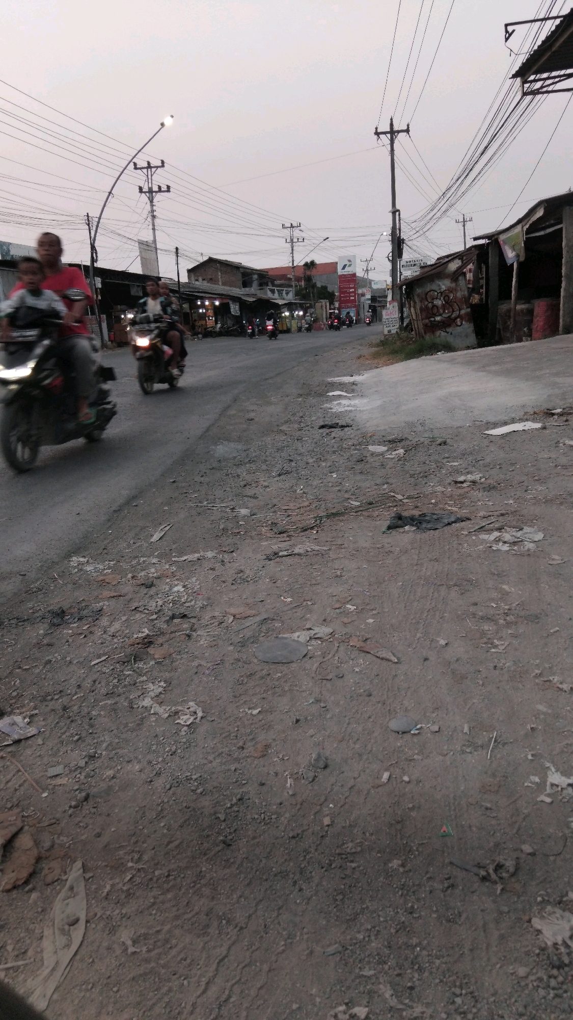 The image shows a street scene in Dukuhturi, Indonesia. In the foreground, the ground is covered in dirt, gravel, and debris. A road runs through the middle ground, with several motorcycles and their riders. The motorcycles are moving in the same direction. On either side of the road, there are buildings that appear to be shops or small businesses. Some of these buildings have awnings or tarps. In the background, there are utility poles with numerous wires crisscrossing overhead. The sky is overcast, suggesting it may be late afternoon or early morning. The overall impression is of a somewhat dusty and busy street in a developing urban or semi-urban area.