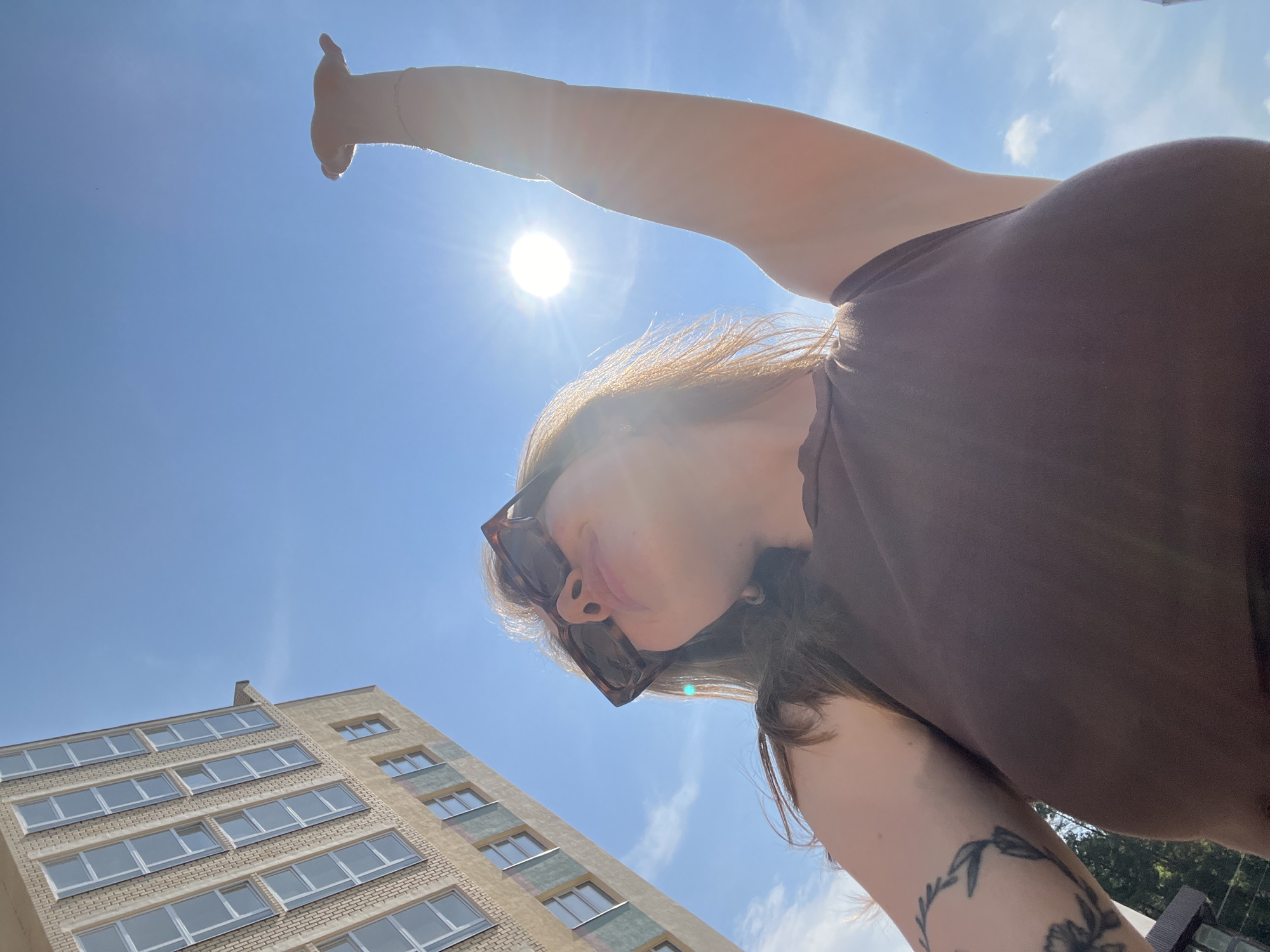 A woman with long, brown hair and sunglasses is looking up at the sky. Her right arm is raised, with her hand outstretched towards the sun. She is wearing a brown top. To her left, a tall, light-colored building with many windows stands against a bright blue sky with a few white clouds. The sun is shining brightly, creating a lens flare in the image. This scene appears to be outdoors in an urban or semi-urban environment, possibly Chaikovsky, Russia, given the architecture.