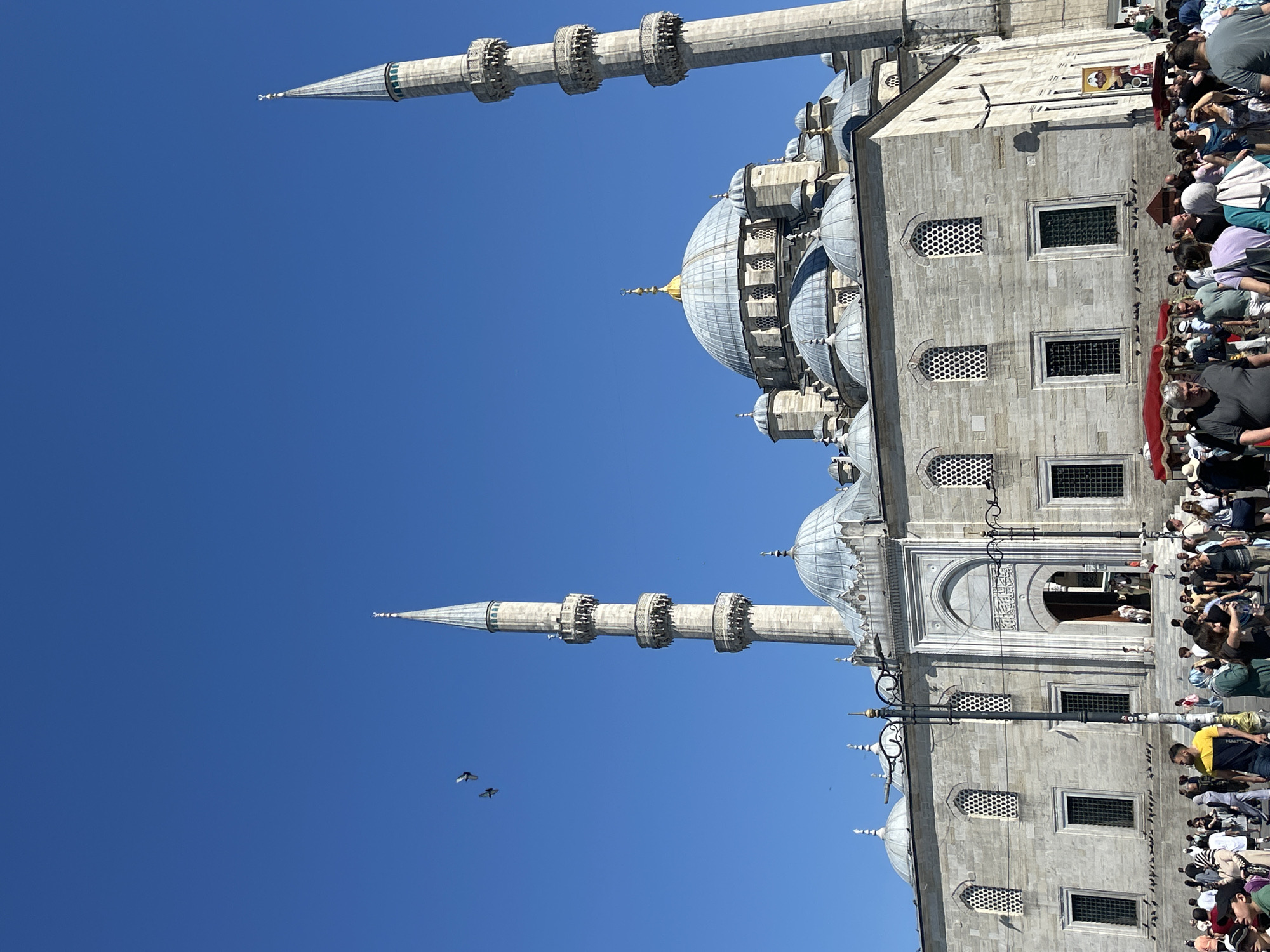 The image shows the Yeni Camii (New Mosque) in the Fatih district of Istanbul, Turkey. The mosque is a large, impressive structure made of stone, featuring multiple domes and two prominent minarets. The sky is clear and blue, indicating a sunny day.
In front of the mosque, a crowd of people is gathered, suggesting a public space or tourist attraction. Some people are walking, while others appear to be standing and observing the mosque. There are also several pigeons in the foreground and in the air around the minarets. Red awnings are visible, possibly indicating food vendors or stalls.
The architecture of the mosque is a key feature, with intricate details on the minarets and domes. The stone facade of the building is visible, along with arched windows and doorways. The overall scene conveys a sense of bustling activity in a historically significant location.