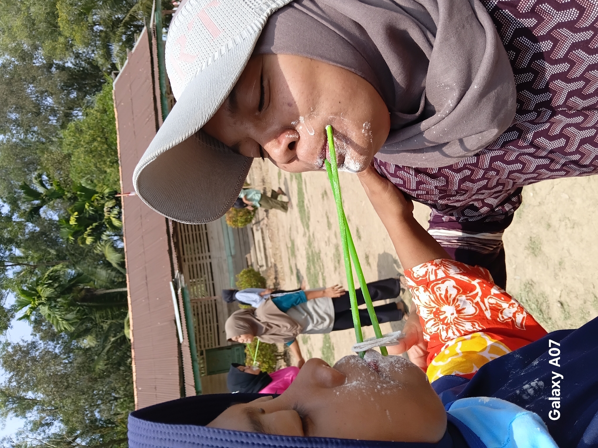 In Margo Rukun, Indonesia, two young women are participating in an outdoor game. The foreground features one woman wearing a blue hijab and a colorful shirt, with a white powdery substance on her face, holding a green straw with her teeth. To her right, another woman, also wearing a hijab and a patterned shirt, is engaged in the same activity, with white powder around her mouth and holding a similar green straw. Both women appear focused on the challenge. In the background, a rustic building with a brown metal roof and open-sided walls is visible, surrounded by trees and vegetation under a clear sky. Other individuals can be seen in the distance, suggesting a community event or gathering. The ground is covered in dirt and sand. The image is marked with "Galaxy A07" in the bottom left corner.