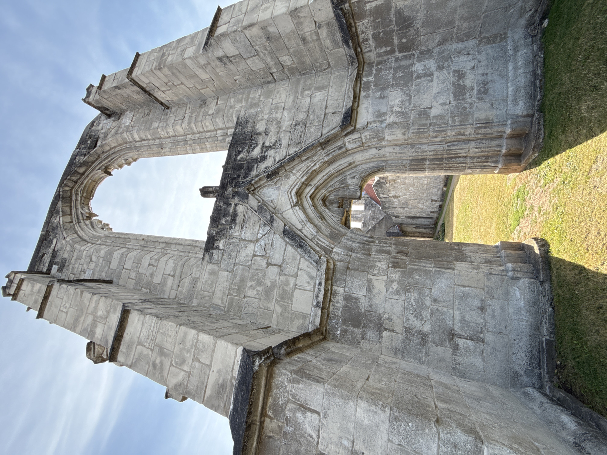 The image shows the ruins of a Gothic-style building, likely a church or abbey, in Walkenried, Germany. The structure is made of weathered stone blocks and features a large, arched opening at the top and a smaller, pointed archway in the foreground. Sunlight illuminates the scene, casting shadows on the grass-covered ground. The sky is blue with some wispy clouds. The ruins are surrounded by a grassy area, suggesting they are part of a historical site or park. No people are visible in the image.