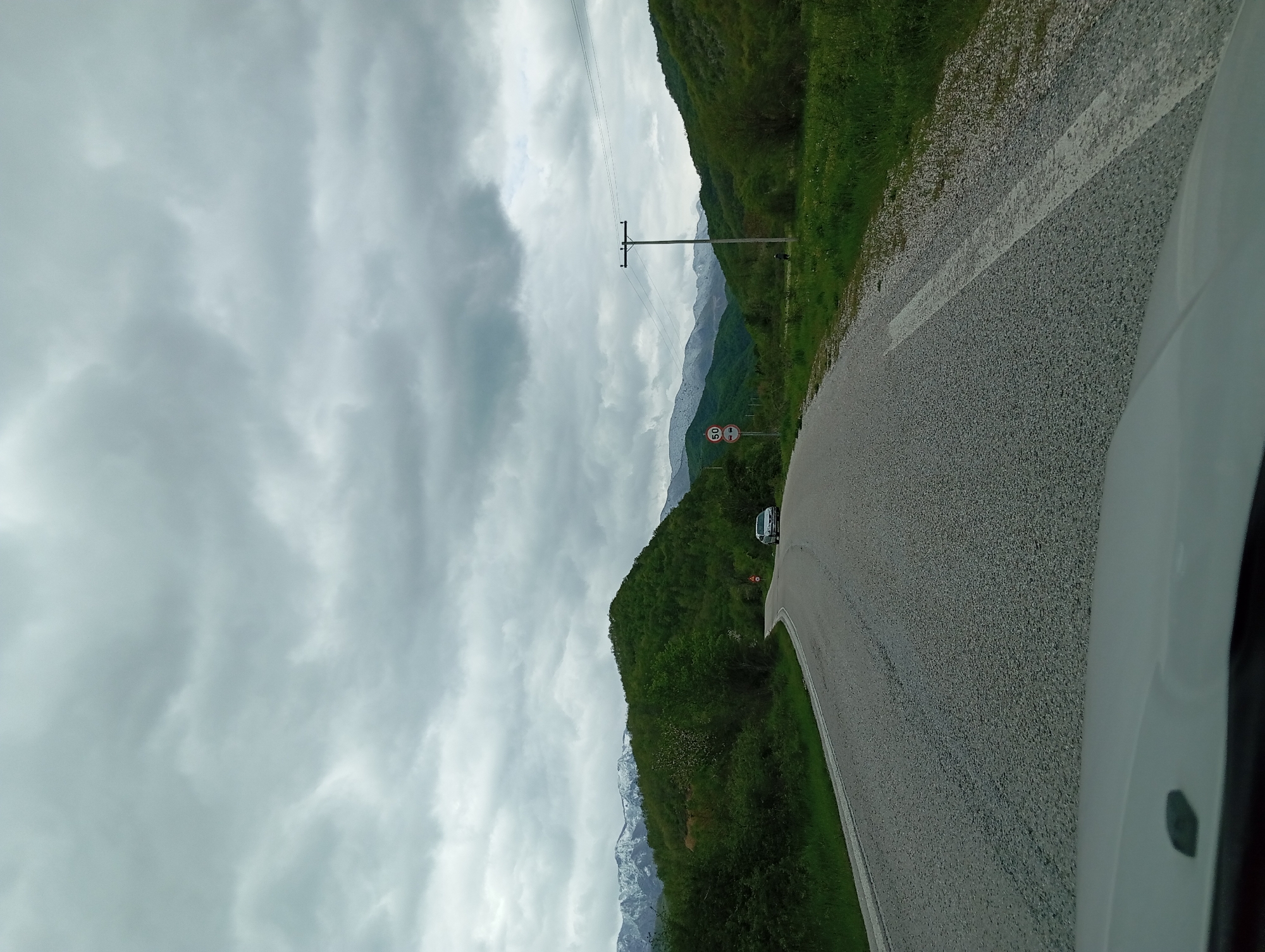 The image depicts a road curving through a mountainous landscape under an overcast sky, potentially located near Melivoia, Greece. A white car is partially visible in the bottom right corner, suggesting the photo was taken from inside a vehicle. The road is flanked by green vegetation and mountains. In the distance, snow-capped peaks are visible through a break in the clouds. A utility pole and wires run parallel to the road. A speed limit sign is also visible. Another vehicle is further down the road.