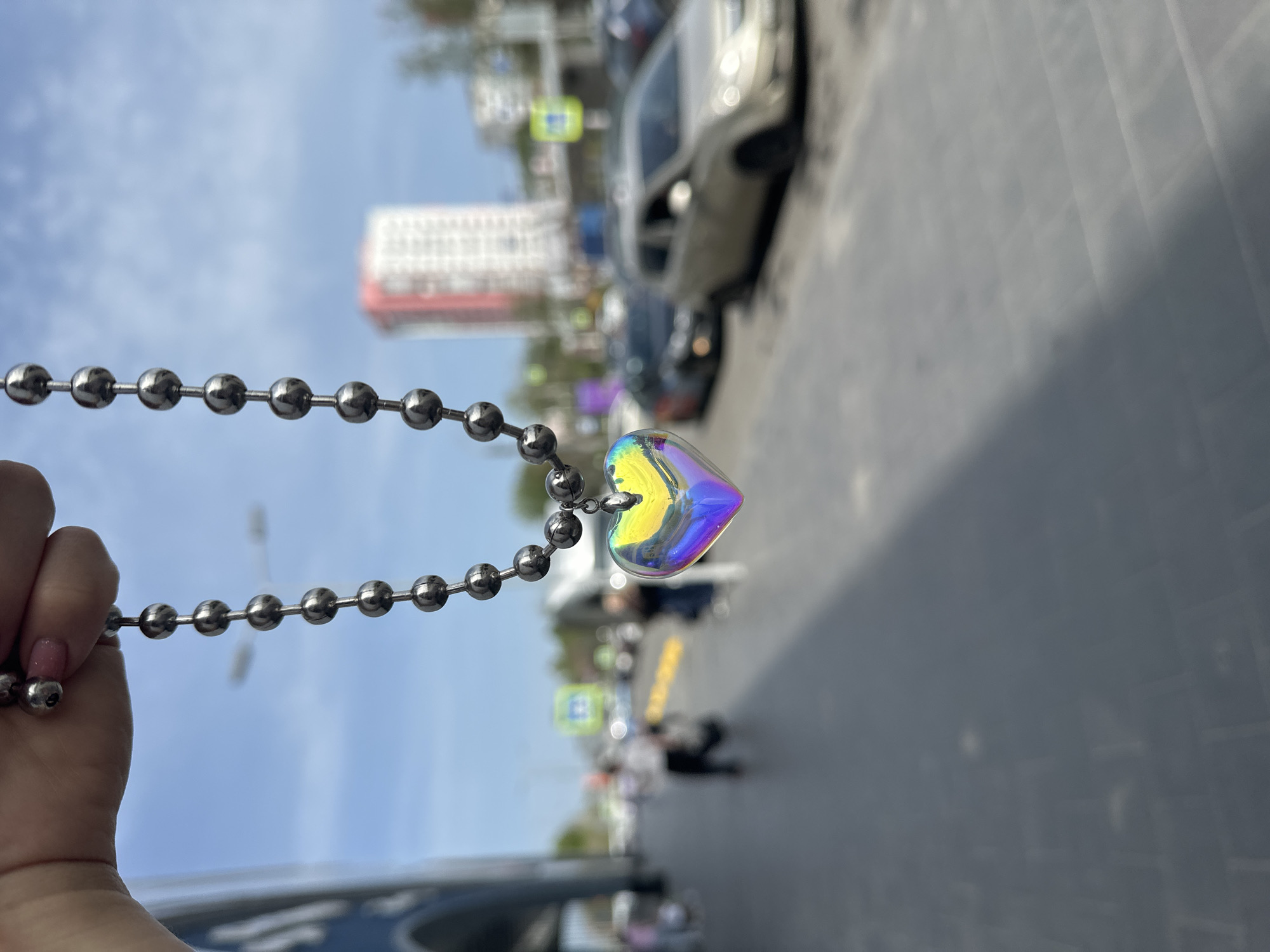 A hand holds a silver ball chain necklace with an iridescent heart pendant in front of an urban street scene in Nizhny Novgorod, Russia. The street contains a building, cars, traffic signs, and several indistinct people walking on the sidewalk. The sky is blue. The image is vertically oriented.