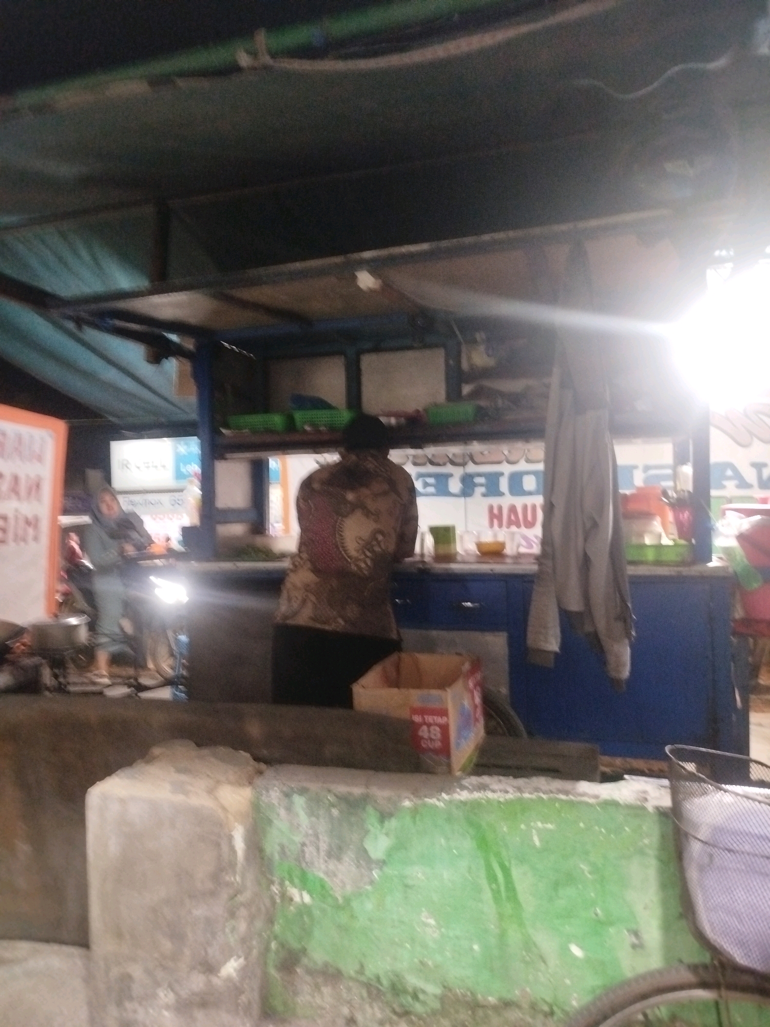 The image depicts a street-side food stall in Burneh, Indonesia. The stall is covered by a dark tarp, with a bright light source to the right of the frame, creating lens flare. In the foreground, there are two concrete blocks, one grey and one painted green, with the green paint peeling. To the right, a bicycle wheel and a basket are partially visible.
In the mid-ground, a person is standing behind a blue counter at the stall, appearing to be engaged in preparing or serving food. They are wearing a patterned shirt. To the left of the stall, another person, dressed in a grey hijab and light-colored clothing, is standing near a motorcycle. The motorcycle's headlight is illuminated, casting light on the ground. A large sign with orange and white lettering is visible on the left. The stall features shelves with green baskets containing items, and the blue counter has drawers.
The overall scene suggests a busy evening at a local food vendor.
