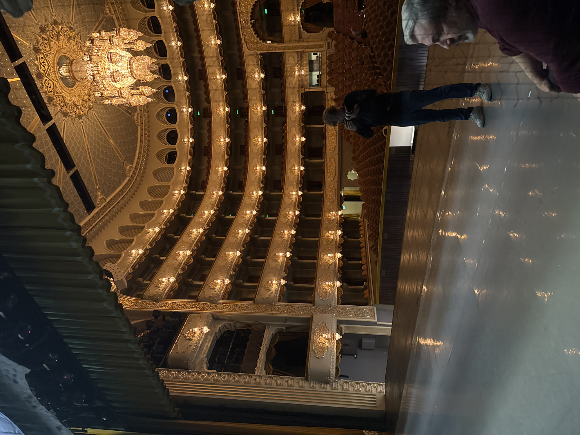The image captures the interior of the Tbilisi Opera and Ballet Theatre in Tbilisi, Georgia. The shot is taken from the stage, looking upwards and outwards towards the multi-tiered balconies and the ornate ceiling adorned with a large chandelier.
Several people are visible. On the right side of the stage, two individuals are standing. One is partially cut off, but appears to be an older man. Another person stands further towards the back, photographing the theatre's interior.
The focus of the image lies on the architectural details of the theatre, including the rows of seats in the balconies, the intricate lighting fixtures, and the elaborate decorative elements of the ceiling. The stage floor is shiny and reflects the theatre's lights.