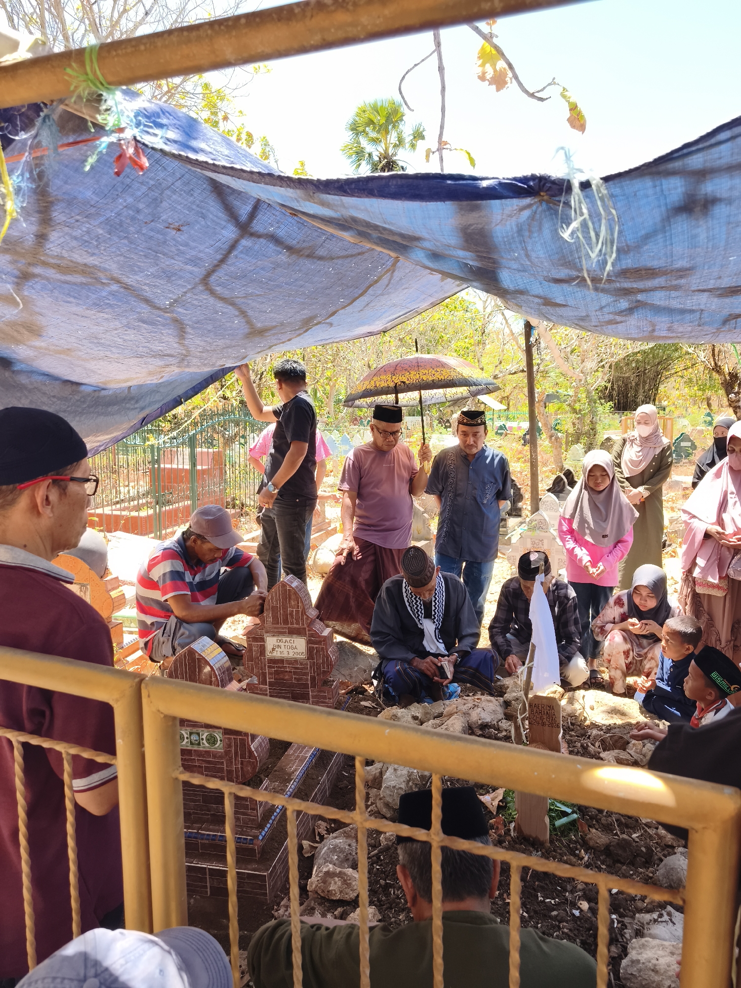 The image depicts a cemetery scene in Tonasa, Indonesia, under a makeshift blue tarp shelter. Several people, both men and women, are gathered around graves. Some are standing, while others are kneeling or sitting. Many are wearing traditional Islamic clothing, including headscarves and caps. A man in the center is holding an umbrella. One grave has a tombstone with an inscription. The environment appears to be outdoors, with trees and bright sunlight visible. A yellow metal fence is in the foreground.