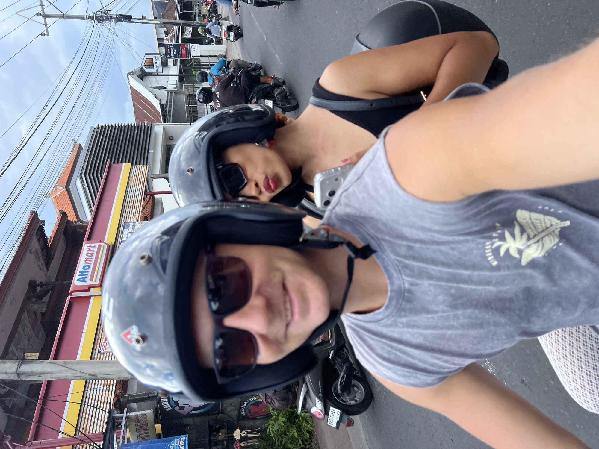 The image shows two people riding a motorcycle in North Kuta, Indonesia. The first person is wearing a grey t-shirt, sunglasses, and a black helmet. The second person is wearing a black top, sunglasses, and a black helmet. They are stopped near an Alfamart store. Other motorcycles are visible on the road. Overhead, there are power lines running between utility poles.