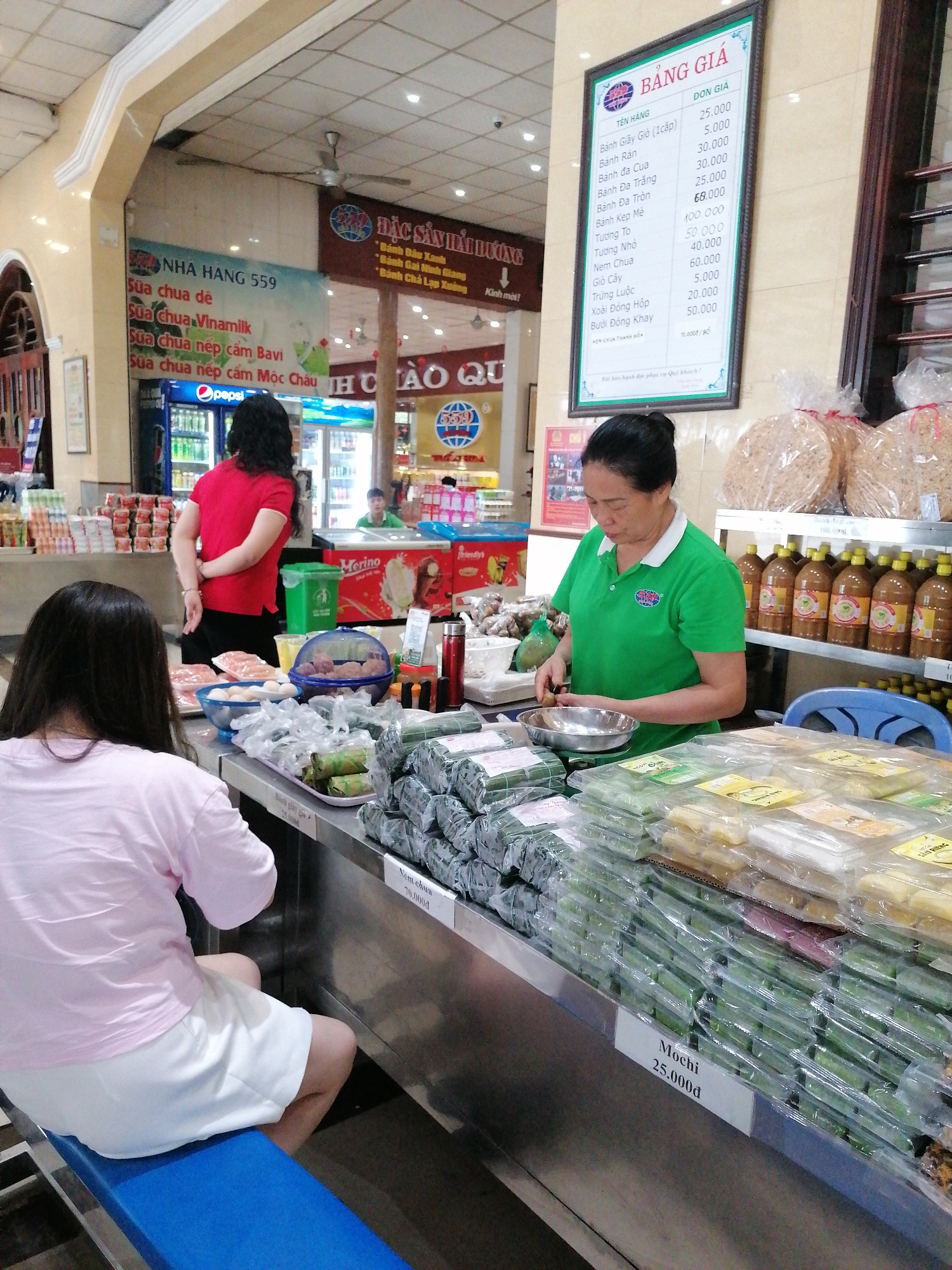 The image shows the interior of a store in Chu Văn An Ward, Vietnam. In the foreground, a woman with long black hair, wearing a pink shirt and white shorts, is seated. Behind her stands a woman in a red shirt with her arms crossed. To the right, an older woman in a green polo shirt prepares food at a counter filled with stacks of packaged goods, including mochi labeled at 25,000₫. A sign displaying a price list hangs above the counter. Shelves behind the counter display bottled products and stacked snacks. In the background, signs for "NHA HANG 559" and "ĐẶC SẢN HẢI DƯƠNG" are visible, along with refrigerators containing beverages and ice cream.