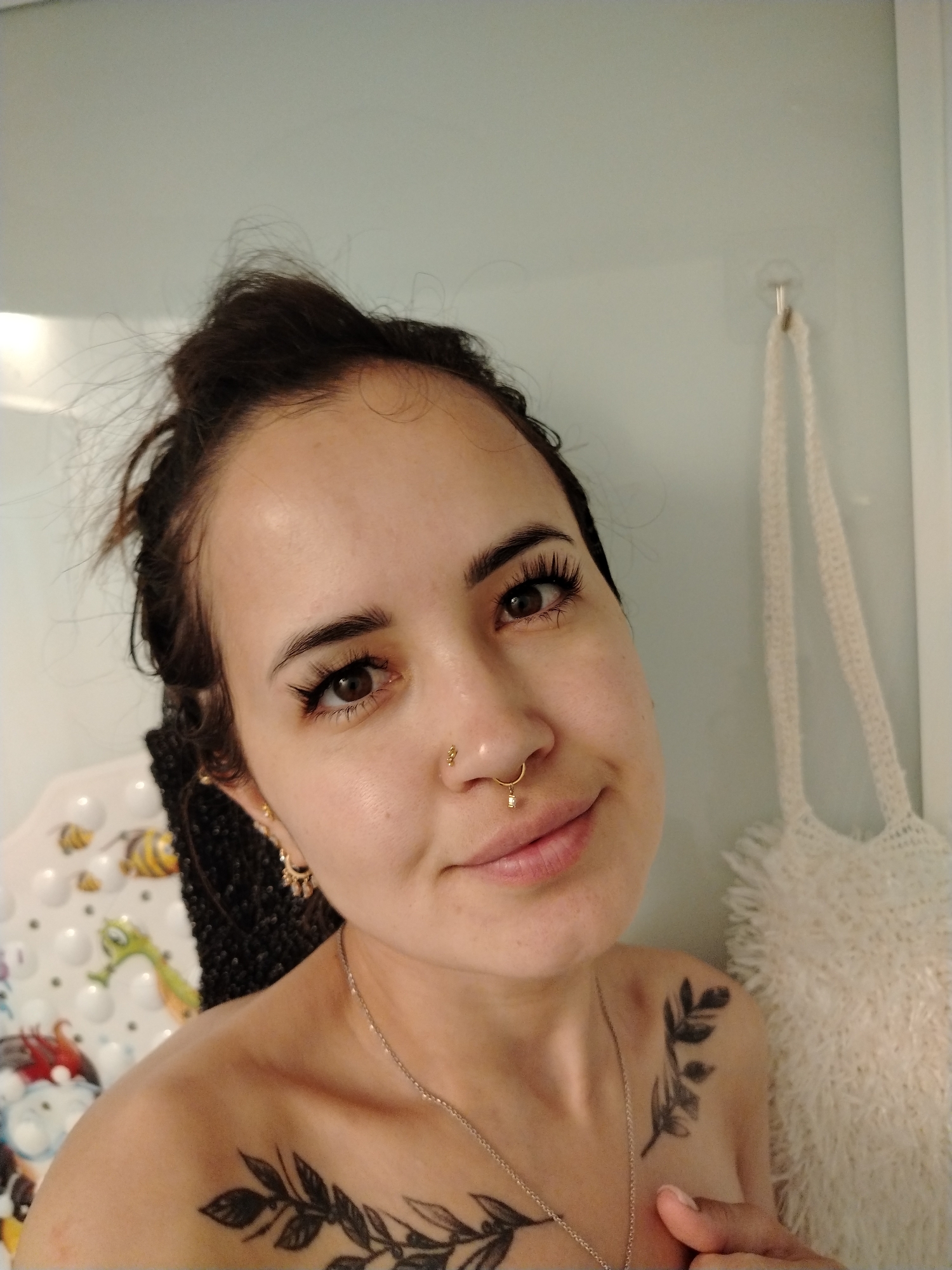 A young woman with dark hair pulled up in a bun, wears a nose ring and a delicate silver necklace. She has on a pair of gold hoop earrings and a septum piercing. A tattoo of branches and leaves is visible on her left shoulder and upper chest. The background appears to be a bathroom, with a shower curtain and a bath mat with cartoon bees and a seahorse. A white crocheted bag hangs on a hook to the right of the frame. There is no indication of the image being taken in Noviy, Russia.