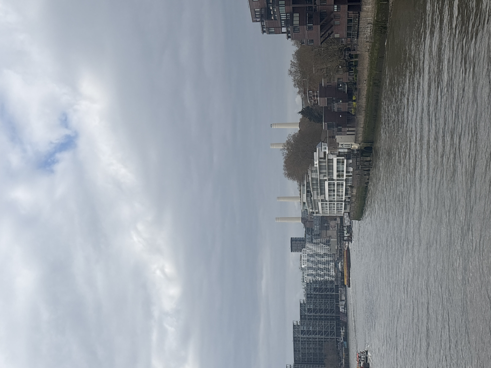 This image depicts the River Thames and the cityscape of London, United Kingdom. The primary feature is the Battersea Power Station, distinguished by its four chimneys, situated along the riverbank. The foreground shows the river's surface, with visible ripples and reflections. Adjacent to the river, a concrete embankment runs alongside the river. Buildings of various architectural styles, including modern residential developments and taller structures, line the riverbank. The sky is overcast. No people are clearly visible.
