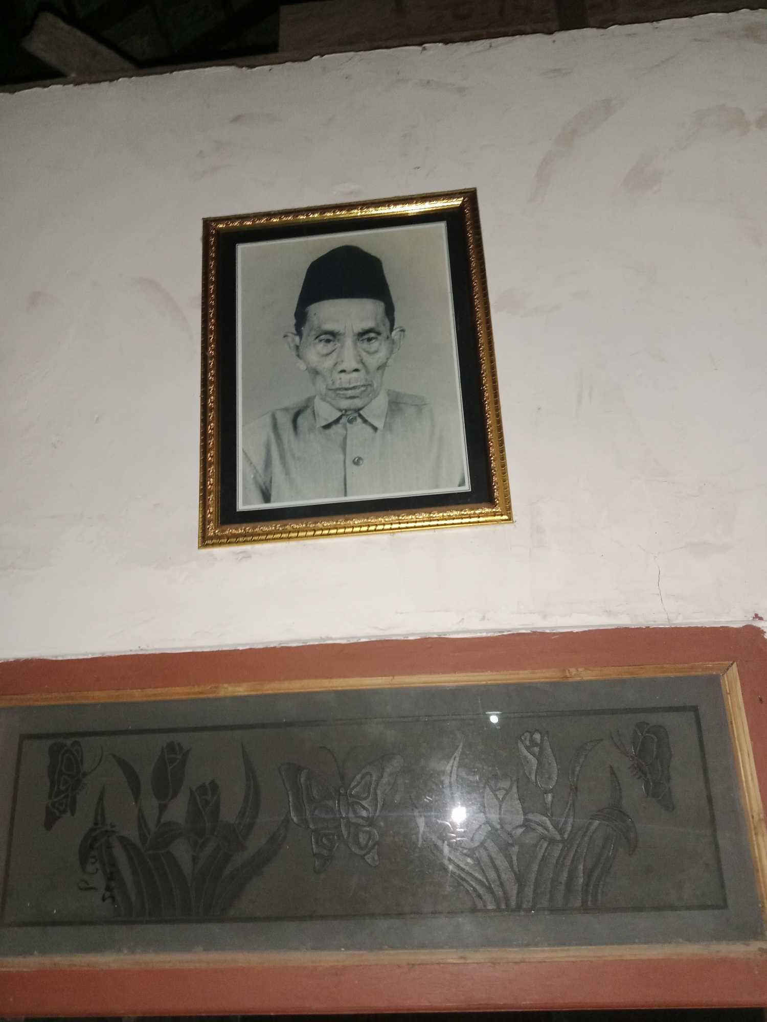 The image is a medium-angle shot of a framed black and white portrait of an elderly man wearing a black cap and a collared shirt. He is looking directly at the camera with a neutral expression. The portrait is mounted on a white wall with some staining and cracks. Below the portrait is another framed artwork, a dark rectangular panel with a relief carving of flowers and butterflies. The framing of both the portrait and the panel is made of wood, with the portrait having a gold-colored ornate frame and the lower artwork having a reddish-brown frame. The scene appears to be indoors, possibly in a home in Ringinrejo, Indonesia, given the context provided. The lighting is somewhat dim, with a reflection visible on the glass of the lower artwork. The ceiling area above the portrait shows a dark, textured surface, possibly wooden beams or roofing.