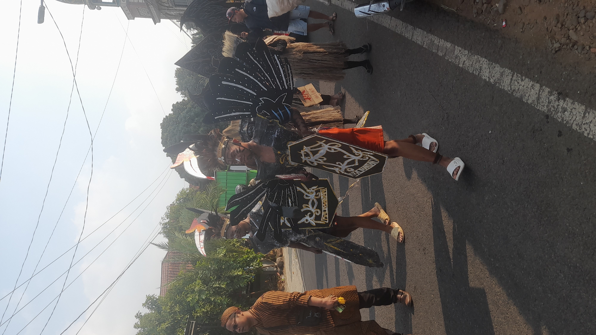 This image depicts a procession or parade in Lemahabang, Indonesia, under a bright, overcast sky. The foreground shows a paved road with a white line marking the edge. Several people are walking on the road. The central figures are participants in traditional or ceremonial attire. Two individuals are prominently featured, wearing dark, feathered headdresses and elaborate, wing-like shoulder adornments in black and white, resembling stylized birds. They also carry decorated shields. One of them wears an orange skirt, and the other wears a dark skirt with yellow and white patterns. To the left, a man in a brown striped, traditional Indonesian shirt and pants walks, holding a yellow object. Further back, other participants are visible, some with straw-like skirts, and onlookers or other participants in casual clothing can be seen on the sides of the road and further down the street. The background reveals trees, buildings with Indonesian architectural elements, including what appears to be a mosque, and utility poles with numerous wires stretching across the sky. The scene is well-lit, suggesting it is daytime, possibly in the morning or late afternoon.