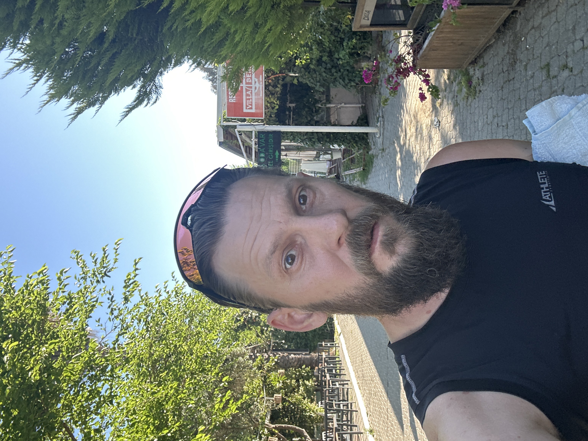 The image is a selfie taken outdoors in Fethiye, Turkey, featuring a man with a full beard and sunglasses pushed up onto his head. He is wearing a black sleeveless top with the text "ATHLETE SPORTSWEAR" visible on his chest. The man has a neutral expression and is looking directly at the camera.
The background reveals a bright, sunny daytime scene with a clear blue sky. Lush green trees, including coniferous-like foliage and broadleaf trees, frame the upper and lower portions of the image. Behind the man, a paved area with cobblestone patterns leads towards buildings.
Several signs are visible in the background. A prominent red sign clearly reads "VILLA BERG," with the word "RESTAURANT" partially visible above it. Below this, a green sign displays text that includes "GİRİŞİDİR," which means "is entrance" in Turkish, suggesting an entrance to a hotel or establishment. Outdoor furniture, such as chairs and tables, can be seen further down the paved area, indicating a cafe or dining setting. Pink bougainvillea flowers grow near wooden planters, adding color to the scene. A light blue towel is draped over the man's right shoulder. The overall bright illumination suggests it is mid-day.