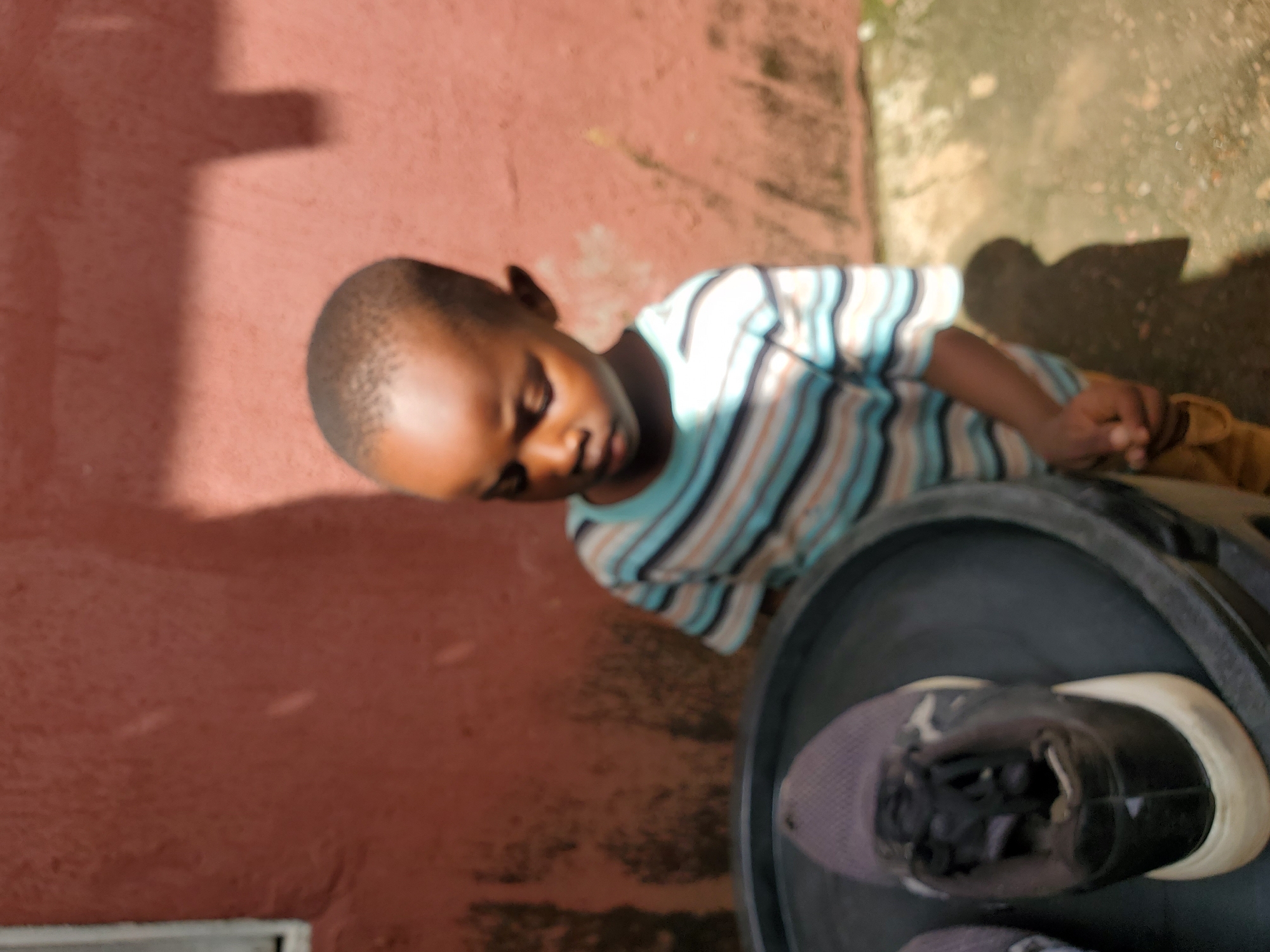 This is a vertical, slightly tilted, medium-shot photo of a young Black boy in Jos, Nigeria. He is wearing a horizontally striped t-shirt with colors of white, blue, and brown. The boy is looking down with his eyes closed, appearing to be resting or in a moment of quiet contemplation.
He is positioned next to a large, dark circular object, which appears to be a bucket or bin. Inside this container, a single sneaker is visible. The sneaker is predominantly black with a light purple mesh upper and a white sole.
The background consists of a reddish-brown textured wall, possibly painted concrete, with some moss or discoloration. Sunlight casts a strong shadow on the wall from an unseen object, suggesting it is daytime, likely the afternoon due to the angle of the shadow. The ground visible to the right of the boy is a mix of dirt and possibly concrete, with some patchy vegetation. The overall setting appears to be an outdoor area, perhaps a yard or alleyway. No visible text is present in the image.