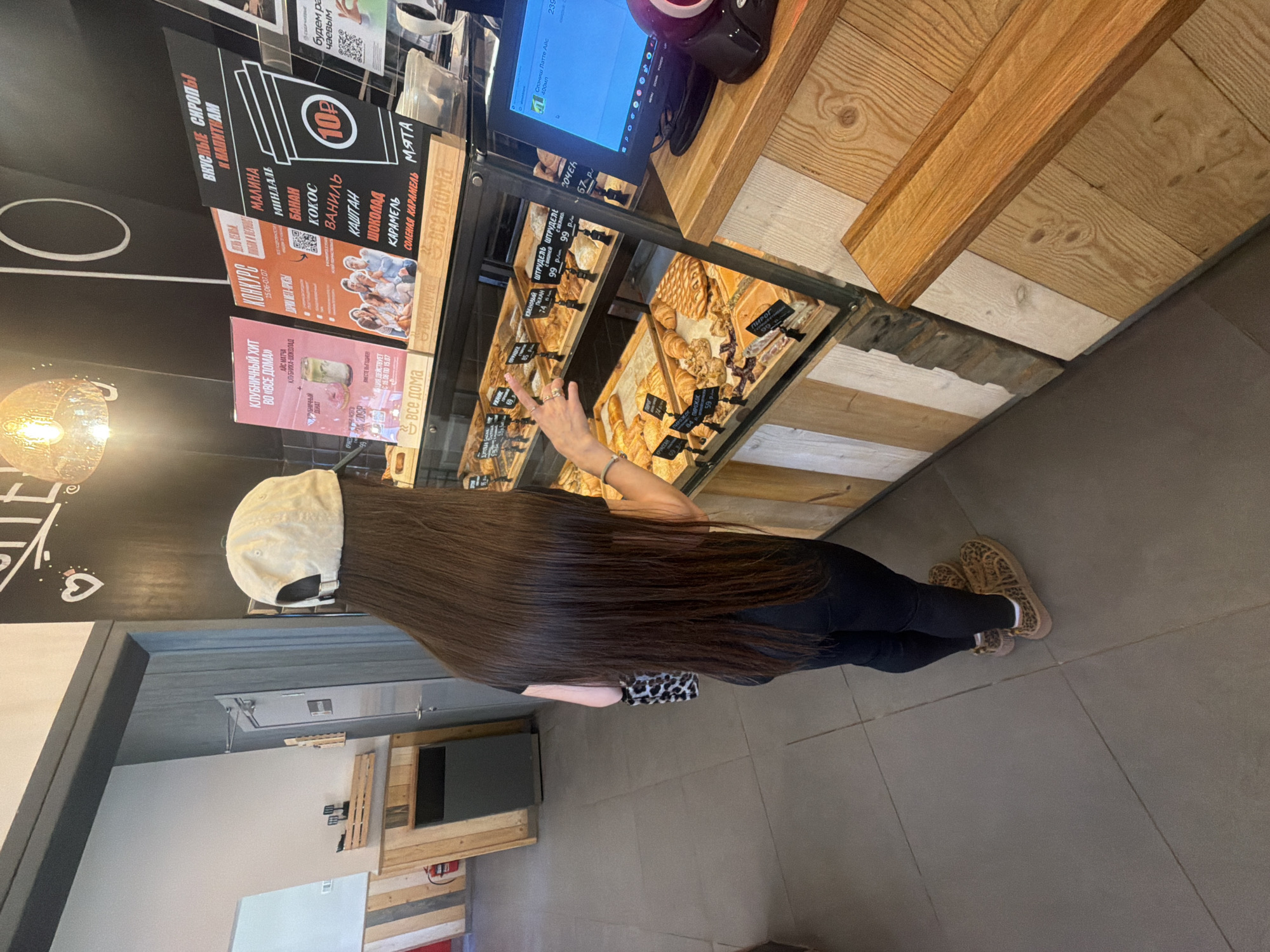 A young woman with long, dark brown hair, wearing a light-colored baseball cap, black leggings, and leopard-print shoes, stands in a bakery in Saint Petersburg, Russia. She is facing away from the camera, looking at a display case filled with pastries. Her left arm is extended, and she appears to be pointing at something in the display. Behind her, a fire extinguisher is visible on a wall. In the foreground, a computer monitor and a webcam are on the counter. The bakery has a modern design with wooden accents and displays of baked goods.