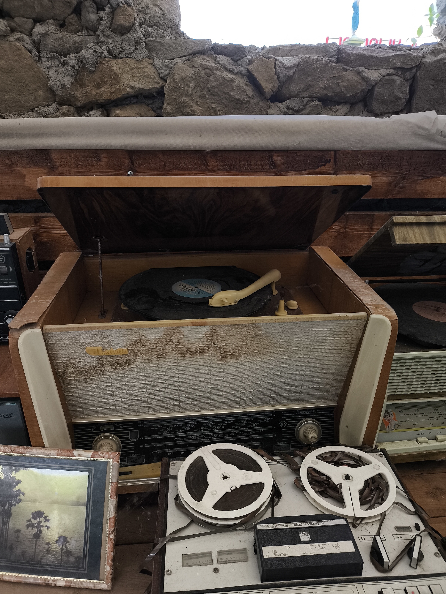 The image depicts an indoor scene with vintage audio equipment and a framed painting, possibly in a storage or display area. The background consists of rough stone walls and wooden planks. In the foreground, a wooden retro record player is open, revealing a vinyl record and a tonearm. To the right of the record player, there are two reel-to-reel tape recorders with spools of tape. A framed painting of a landscape with palm trees and water is visible on the left. The overall impression is one of a collection of old electronic devices and decorative items. There are no people visible in the image. The location is identified as Borgustanskaya, Russia.