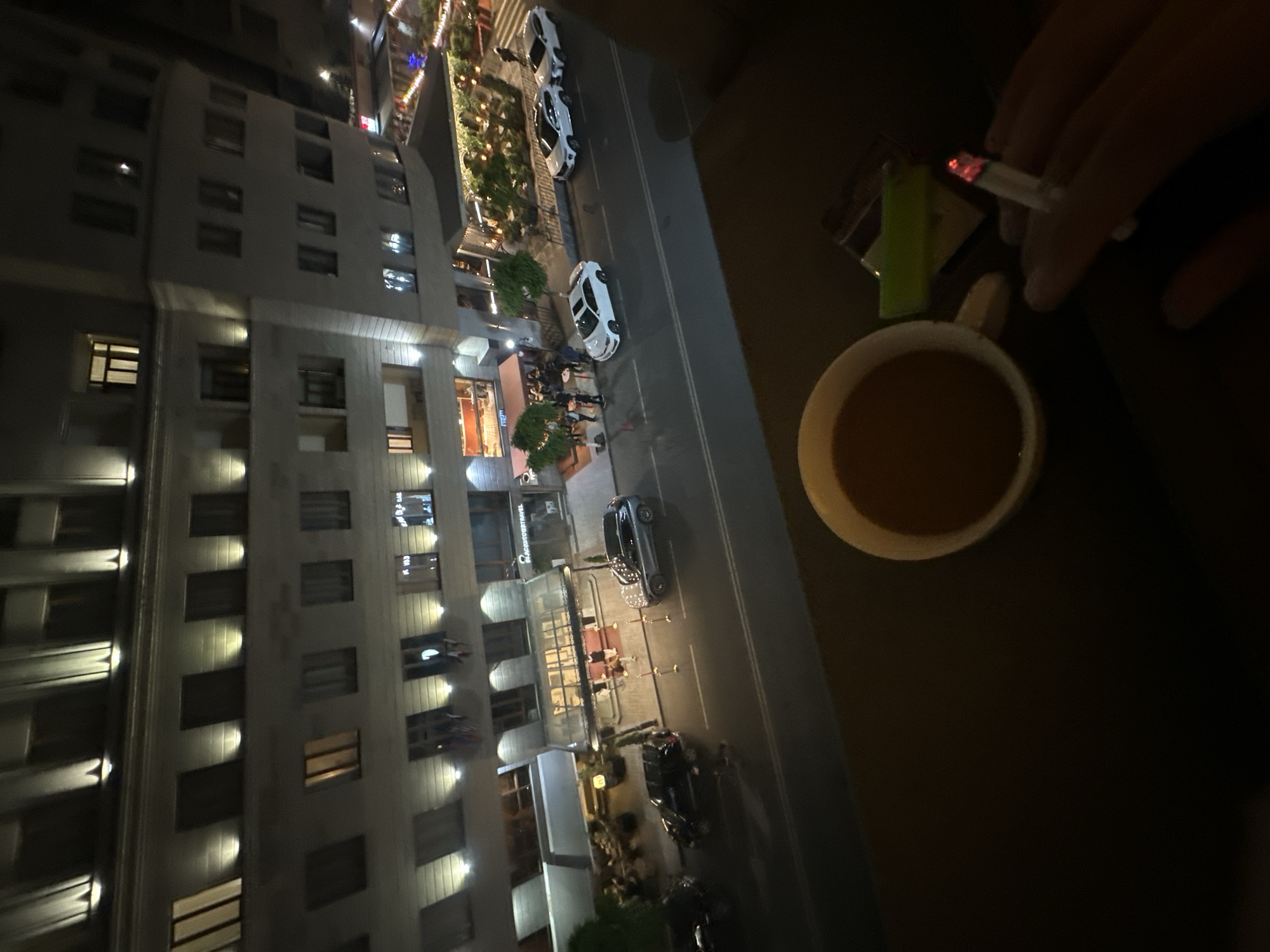 The image depicts a nighttime scene with a high-angle view of a cityscape, likely taken from a balcony or high-rise building. In the foreground, there's a mug of coffee and a hand holding a lit cigarette. A lighter is next to the mug.
The cityscape below shows a building with many windows and street lighting. There are parked cars and pedestrians on the street.
Based on the architectural style and the street layout, the location is identified as Yerevan, Armenia.