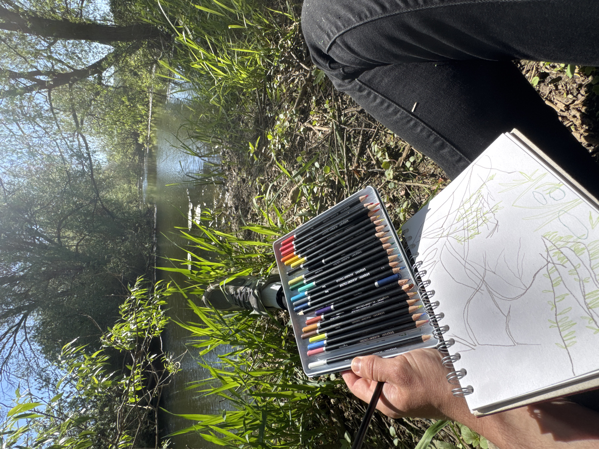 The image captures a first-person perspective of an individual engaged in plein air sketching beside a river or canal in Toll Bar, United Kingdom. In the foreground, a person's hand holds a dark pencil over a white, spiral-bound sketchbook, which displays an initial drawing of green leaves and brown branches. Adjacent to the sketchbook, an open metal tin case holds a collection of "DERWENT ACADEMY COLOUR" pencils, neatly arranged in a spectrum of vibrant colors.
The person is wearing black trousers and grey shoes, suggesting they are seated on the earthy, grass-covered bank. The setting is rich with natural elements: lush green grasses and foliage frame the foreground, leading to a calm body of water that reflects the bright, clear sky and distant trees. Sunlight filters through the dense tree canopy, creating shimmering reflections on the water's surface and highlighting the vibrant greens of the surrounding vegetation. The bright lighting indicates a sunny day, likely late morning or early afternoon, contributing to a serene atmosphere of artistic creation in nature.