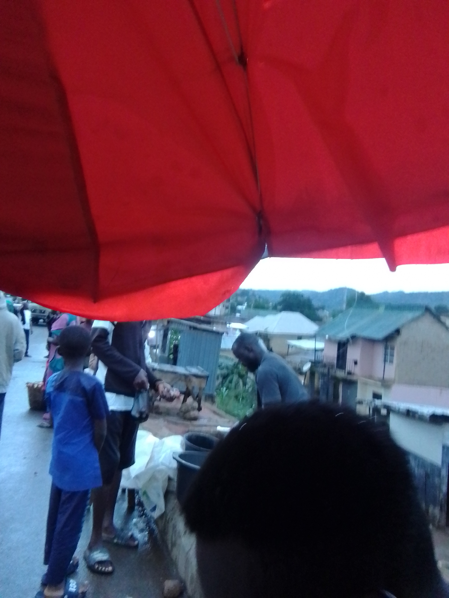 A low-angle, slightly blurred image captures a street scene in Gwong, Nigeria, under a large red umbrella. In the foreground, the dark, rounded shape of a person's head and shoulders obscures the lower right portion of the frame. To the left, a young boy in a bright blue shirt and dark pants stands facing away from the camera, looking towards the center. To his right, a person wearing a white undershirt and dark shorts is partially visible, interacting with a table-like structure. Behind them, another person in a dark shirt is seen in profile, their head tilted downwards. In the background, several buildings with pitched roofs are visible, along with a hazy, mountainous landscape under an overcast sky. The ground appears to be wet, suggesting recent rain or a damp surface. The overall atmosphere is that of a busy, outdoor market or gathering.
