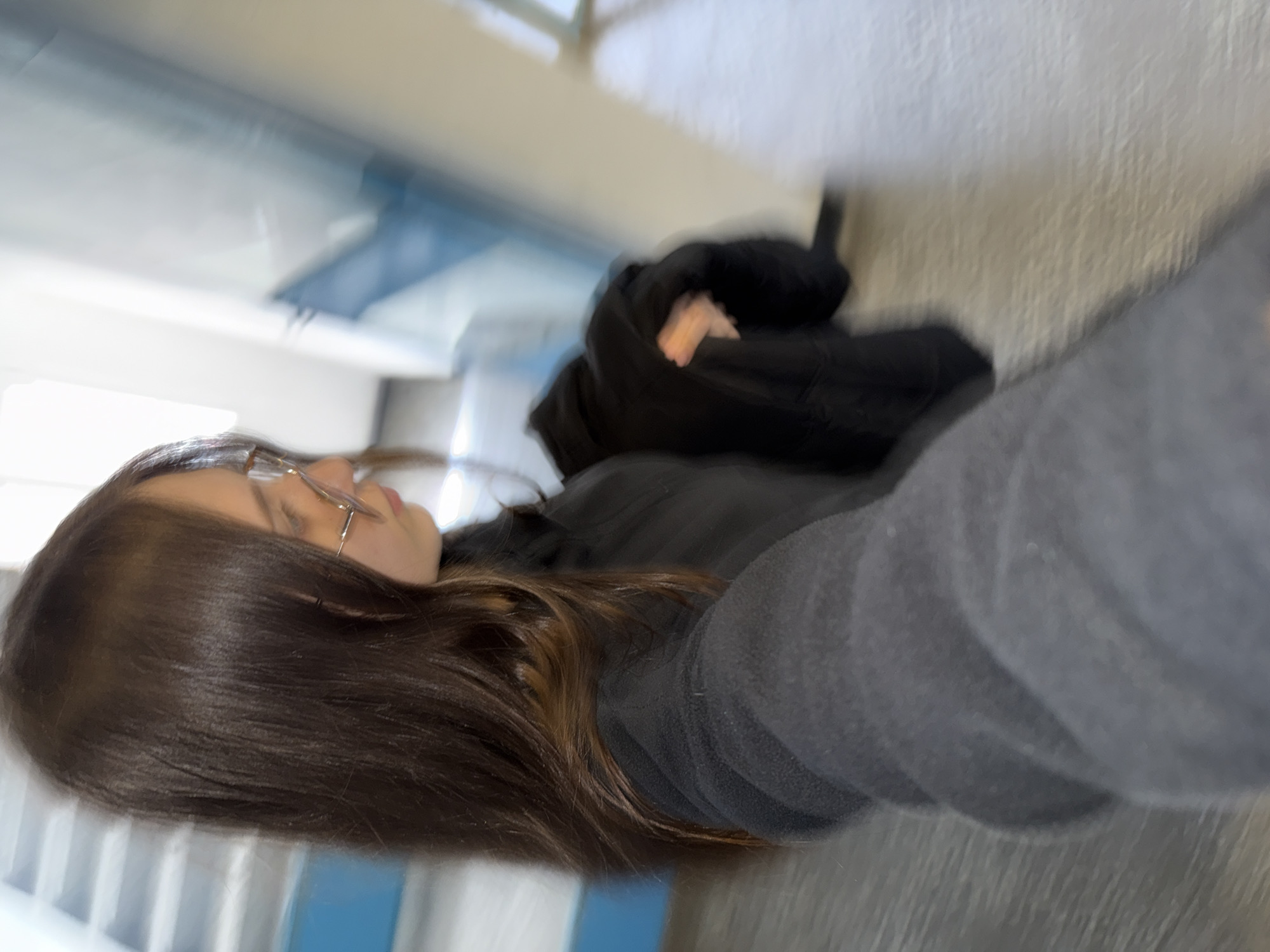 The image displays a person, likely female, in a close-up, motion-blurred shot. The subject, oriented vertically in the frame, has long, straight, dark brown hair flowing over her shoulders. She wears glasses with thin, light-colored frames and dark clothing, including what appears to be a black top or jacket and a dark gray fleece sleeve extending towards the camera.
Her face is visible in profile, looking slightly upwards and to the left. Her expression appears neutral or contemplative, though the blur makes precise details unclear. The background suggests an indoor environment, possibly a hallway or stairwell, with light-colored walls and hints of blue architectural elements or railings further back. The lighting appears bright, indicating a well-lit interior.
The overall blurriness of the image suggests motion during capture, possibly a quick selfie or action shot. There is no visible text. The setting is an indoor location within Sofia, Bulgaria.
