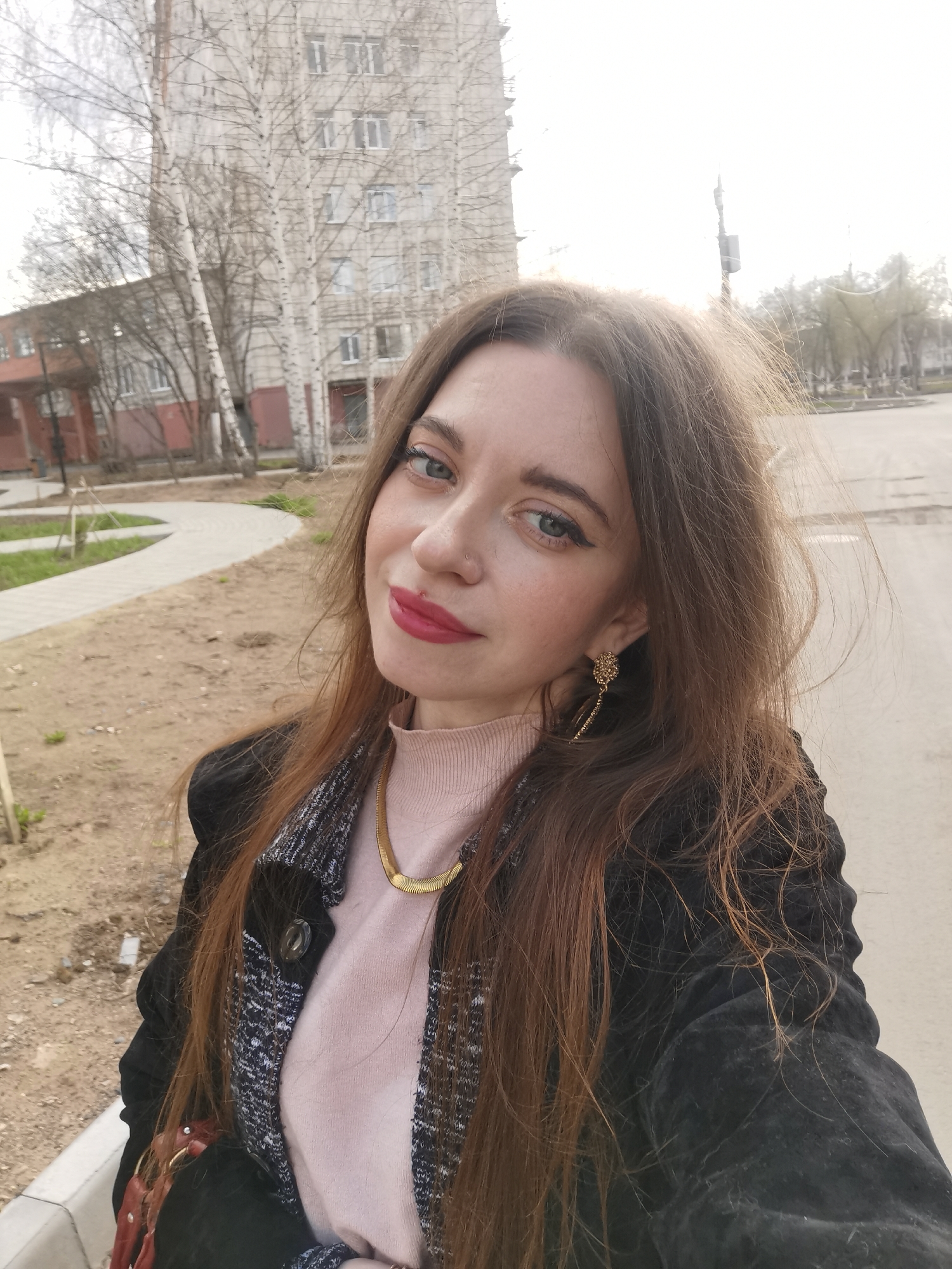 A young woman is depicted in a close-up selfie taken outdoors. She has long, straight light brown hair, blue eyes accented with dark eyeliner, and vibrant red lipstick. A small silver stud piercing is visible on her nose. She is wearing a light pink turtleneck sweater, a prominent gold snake chain necklace, and a dark jacket with a textured grey and black patterned lapel. A decorative gold earring with a dangling chain design adorns her right ear. Her expression is calm and pleasant, with a slight smile as she looks directly at the camera.
The background suggests an urban or suburban setting. To the left, a paved sidewalk winds through an area of dry grass and sparse dirt, featuring several bare deciduous trees, likely birches, whose branches are silhouetted against a bright sky. Behind these trees stands a tall, light-colored building with multiple windows. Another building with a reddish-brown facade is partially visible further left. To the right, an asphalt road stretches into the distance, bordered by more bare trees. The lighting is bright and somewhat diffused, indicating either an overcast day or a sunny day with hazy conditions, and the absence of leaves on the trees suggests late winter or early spring. No text is visible in the image.