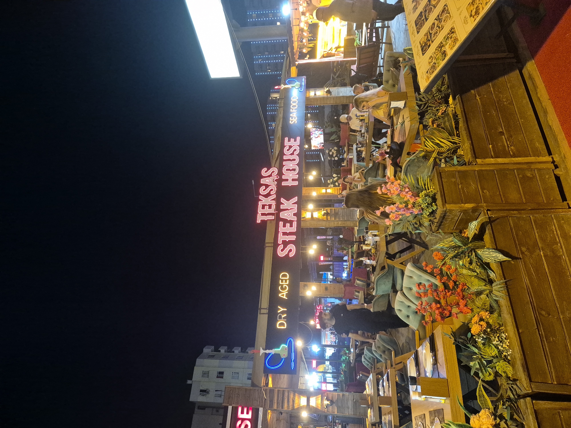 The image depicts a brightly lit restaurant at night, with neon signs and string lights illuminating the outdoor seating area. Several people are visible, seated at tables, appearing to be dining or socializing. The restaurant's signage prominently features "TEKSAS STEAK HOUSE" in large, glowing letters, accompanied by "DRY AGED" and "SEAFOOD." The setting is outdoors, with wooden structures, planters filled with vibrant flowers and greenery, and comfortable seating arranged for patrons. The overall atmosphere suggests a lively and inviting dining establishment. The time of day is clearly night, indicated by the darkness of the sky and the extensive use of artificial lighting. There are no explicit weather cues, but the scene appears to be well-lit and active, suggesting pleasant conditions for outdoor dining. The presence of people engaged in dining and conversation implies a social gathering or a typical evening at a restaurant. The image is captured from a slightly elevated perspective, looking down and across the seating area.